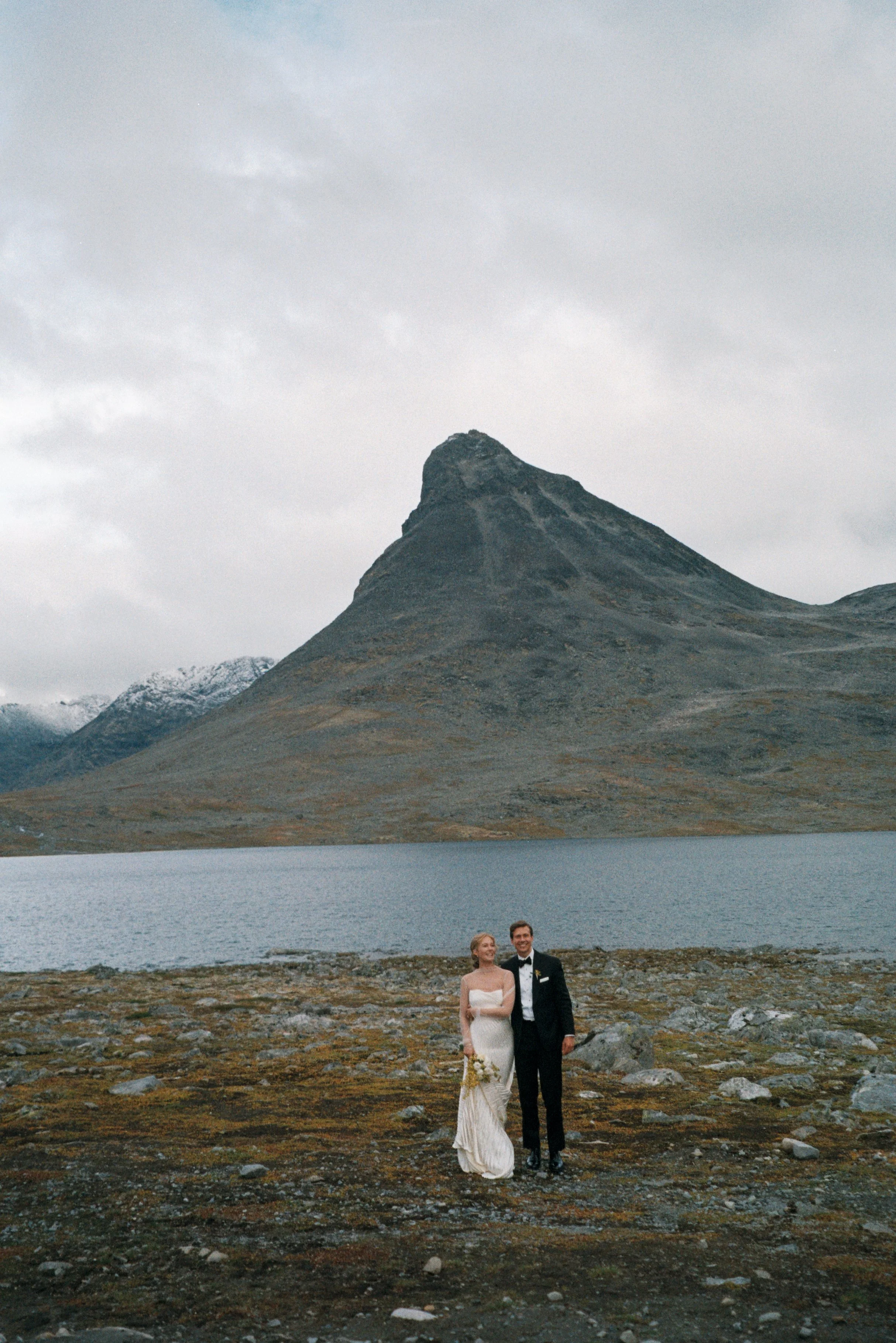 A bride and groom in wedding attire standing by a lake with a mountain in the background.