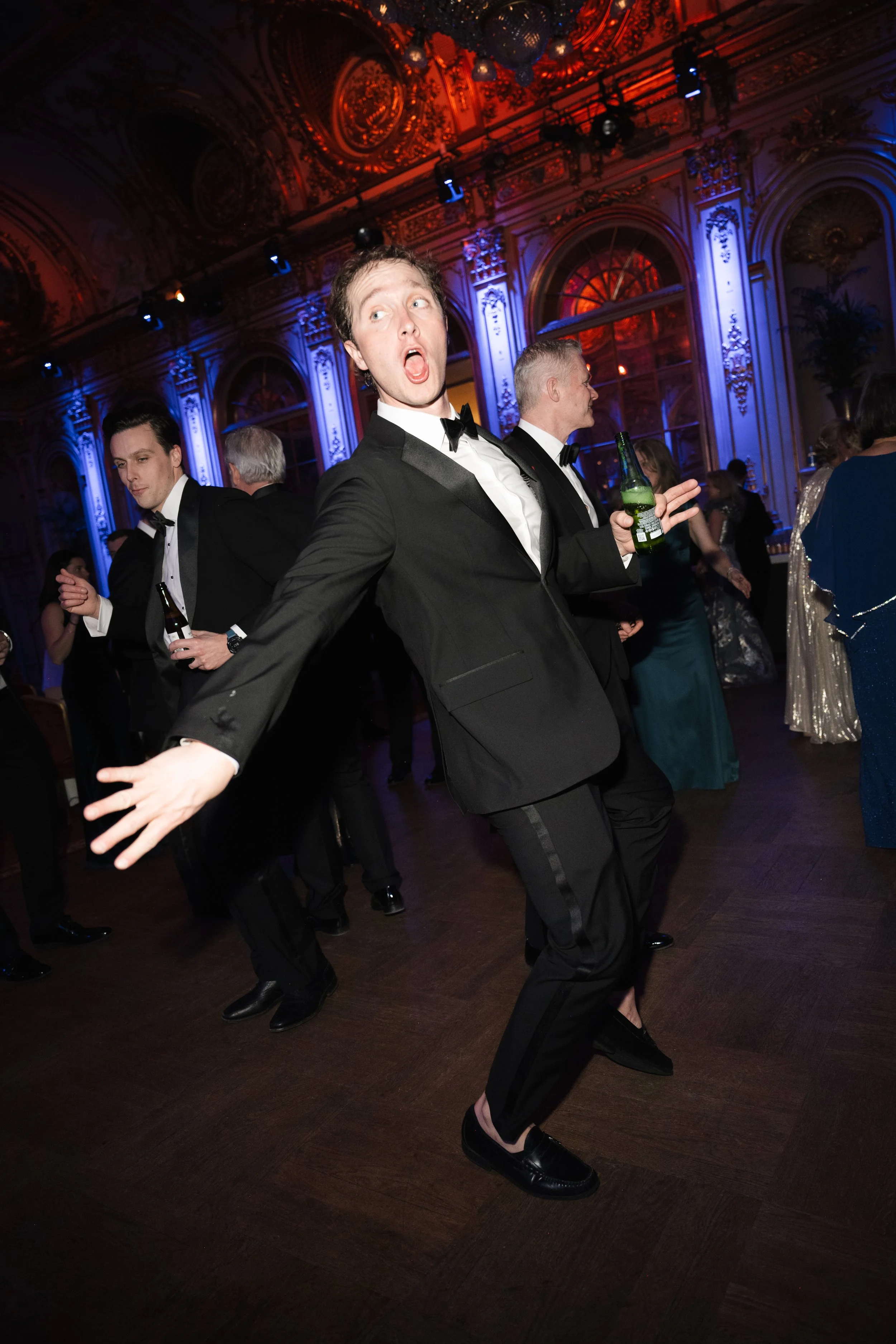 People dancing and socializing at a formal event in an ornate ballroom with blue and purple lighting.