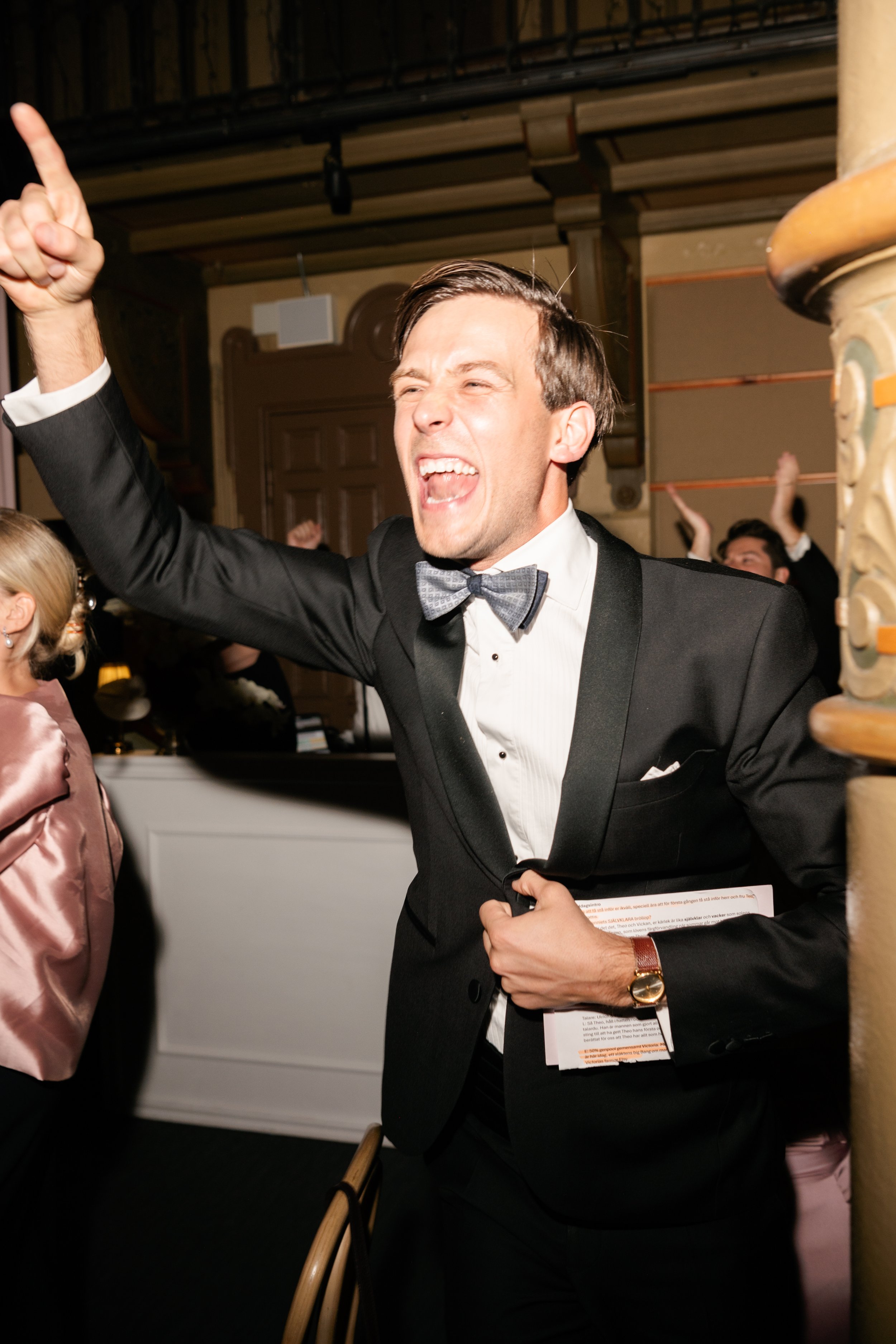A man in a tuxedo with a bow tie passionately singing or shouting at a lively event.
