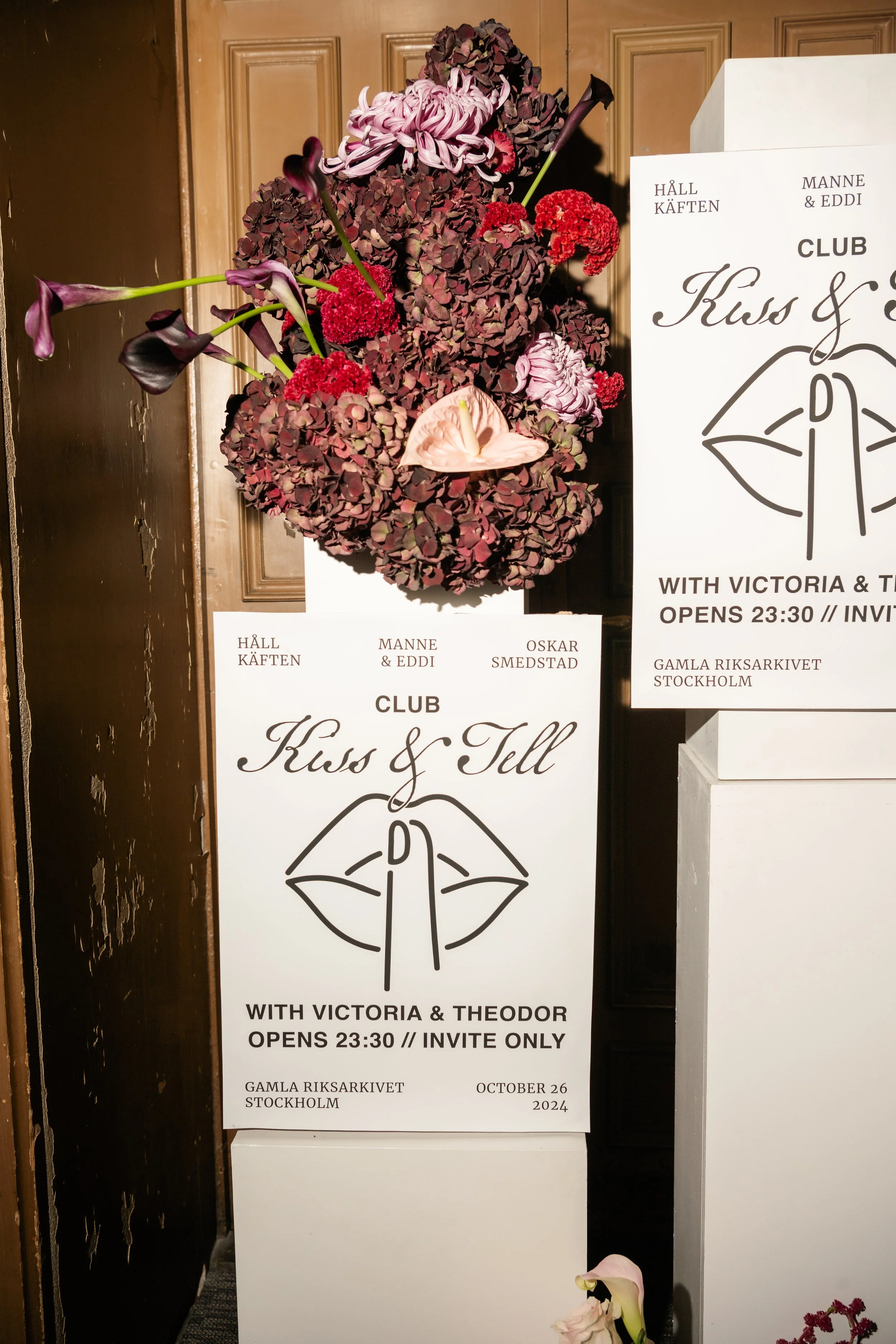 A floral arrangement with pink and dark purple hydrangeas, calla lilies, and red flowers, topped with orchids, next to two white signs with black text and a lip illustration, advertising an exclusive event at the Gamla Riksarkivet in Stockholm on Oct