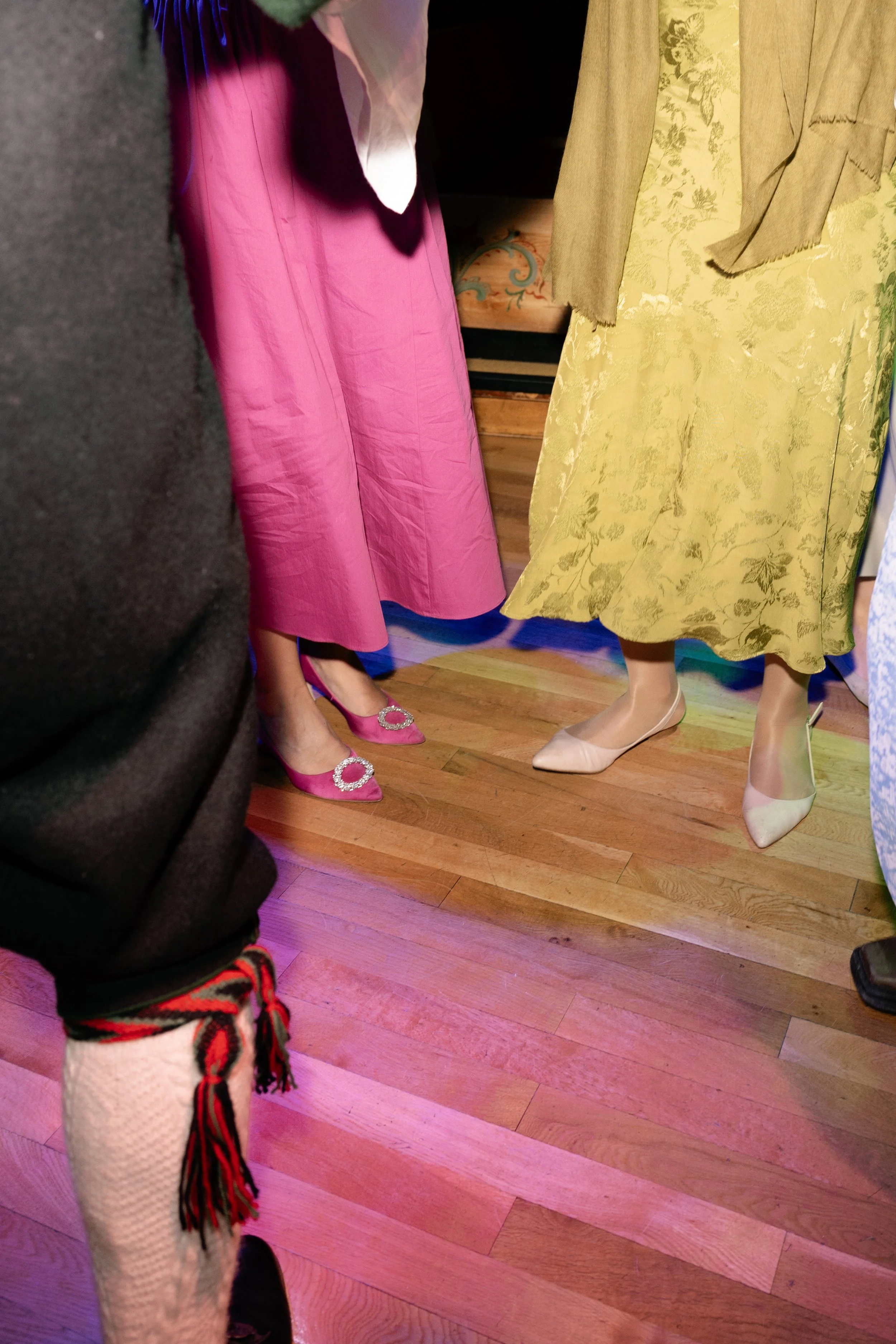 The image shows the lower halves of three people. One person is wearing bright pink dress shoes with emblems, another is in beige heels with a yellow dress, and a third person is wearing black pants with red and white patterned socks.