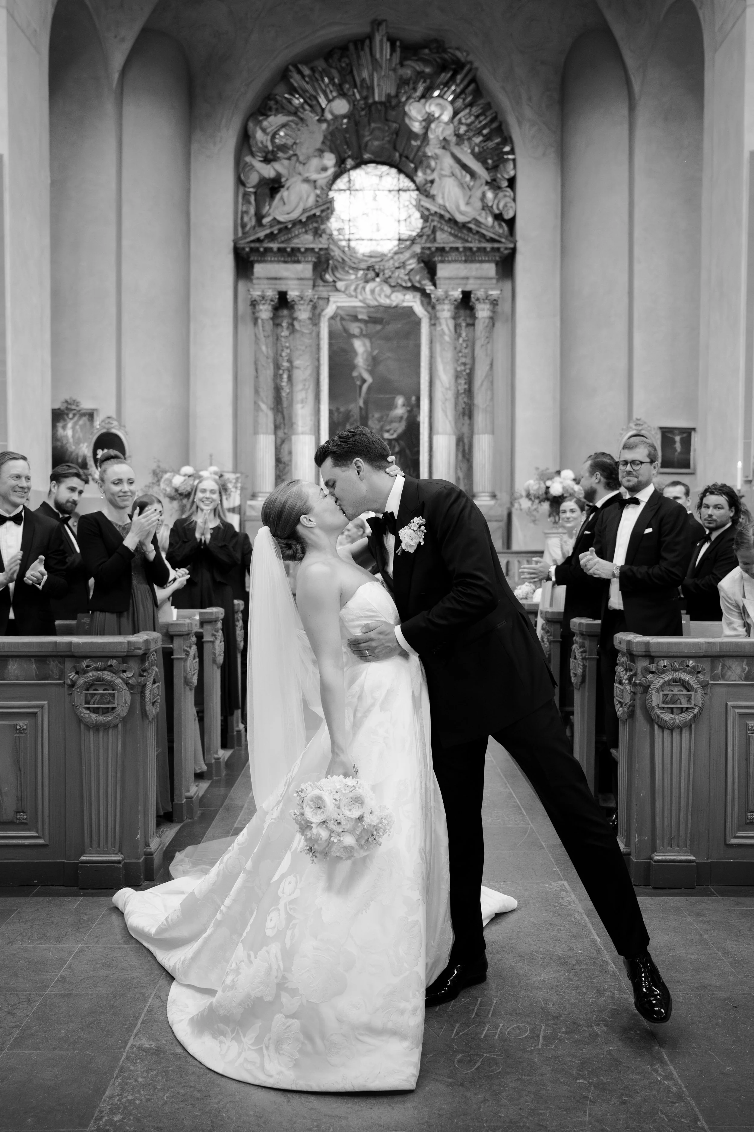 A black and white photo of a newlywed couple kissing at the altar inside a church, with guests clapping and smiling in the background.