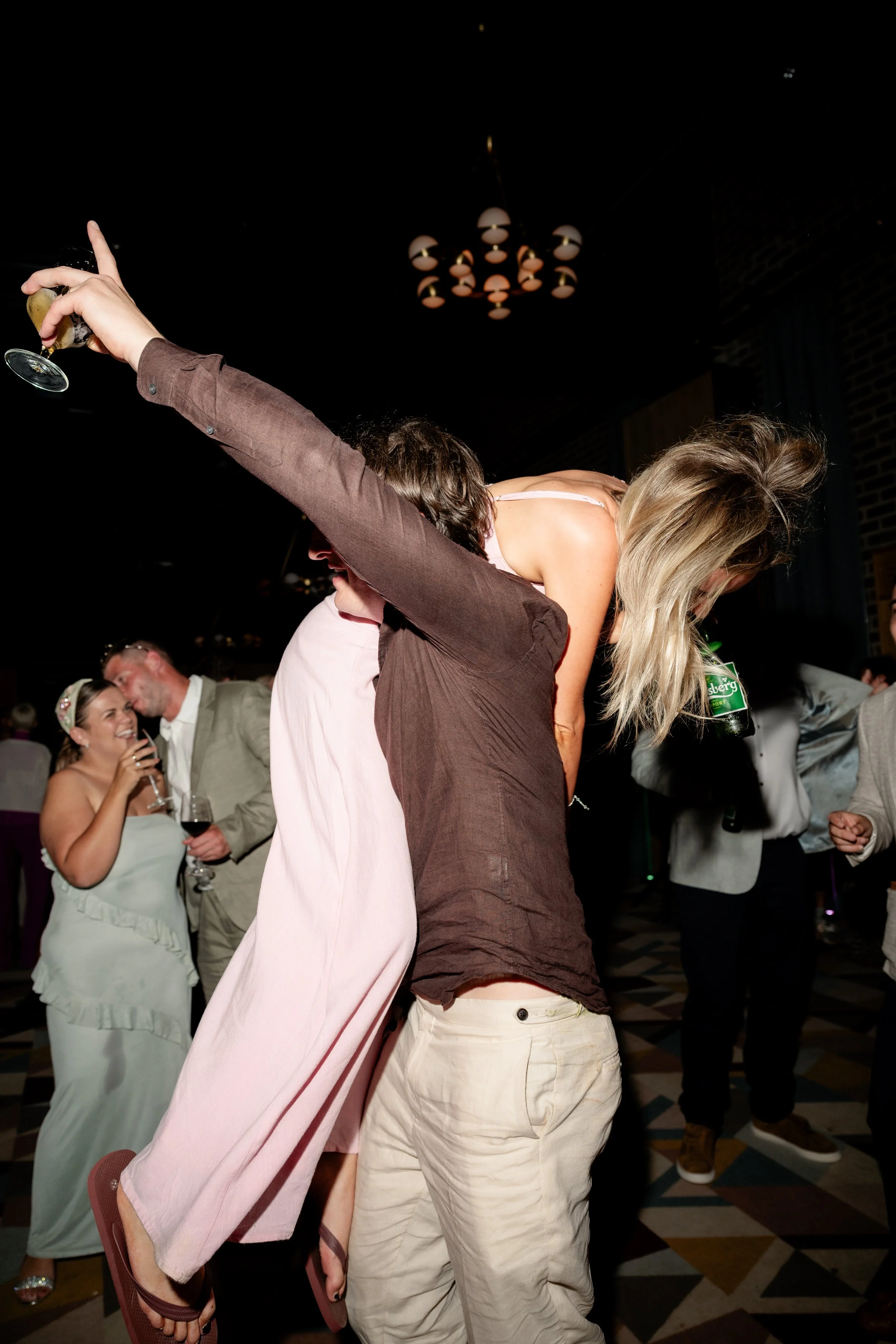 People celebrating at a party with one person giving a piggyback ride to another, who is holding a beer bottle and a glass of wine.
