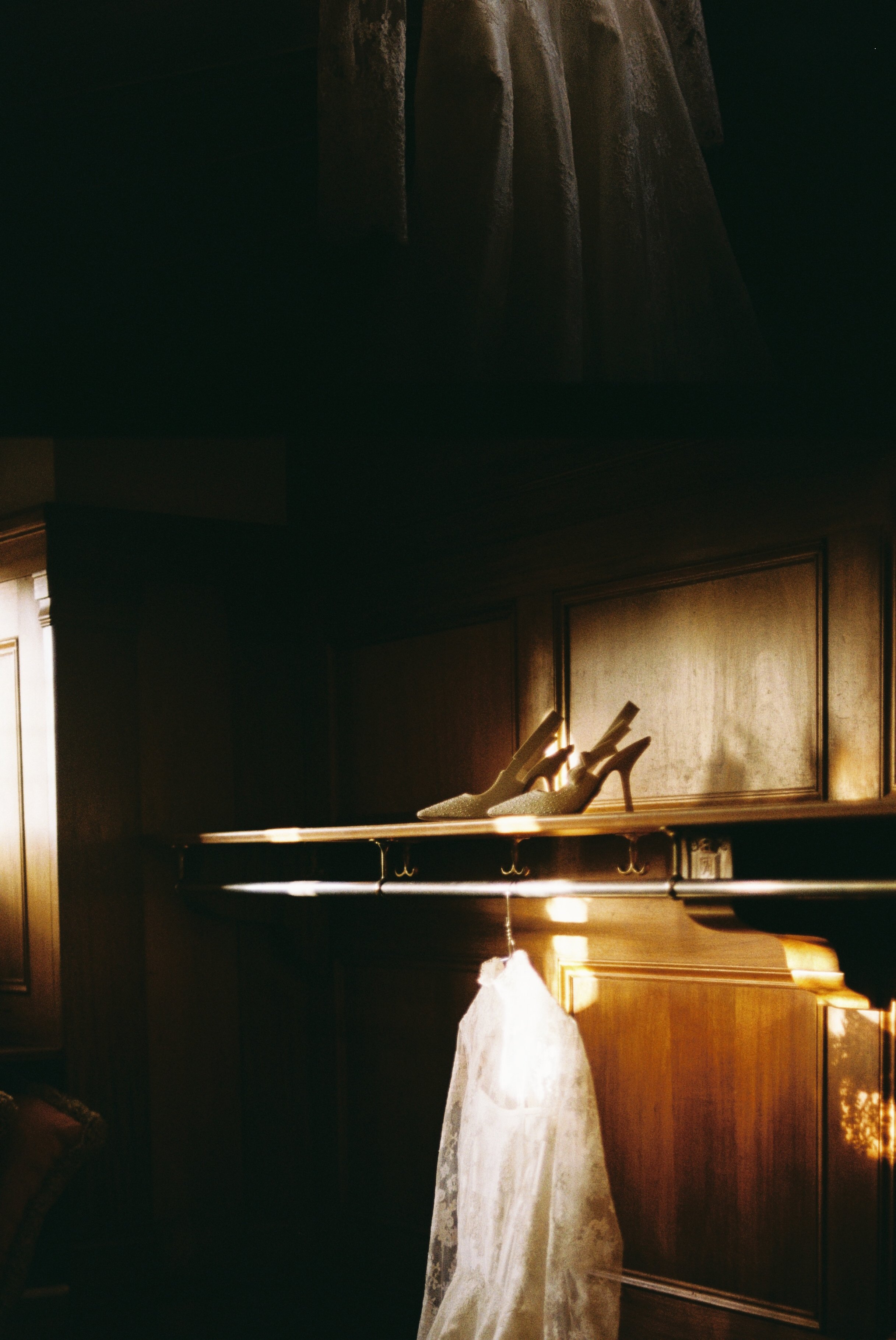 A pair of elegant high-heeled shoes displayed on a wooden shelf, with sunlight casting warm glow and shadows on a lace wedding dress hanging below.