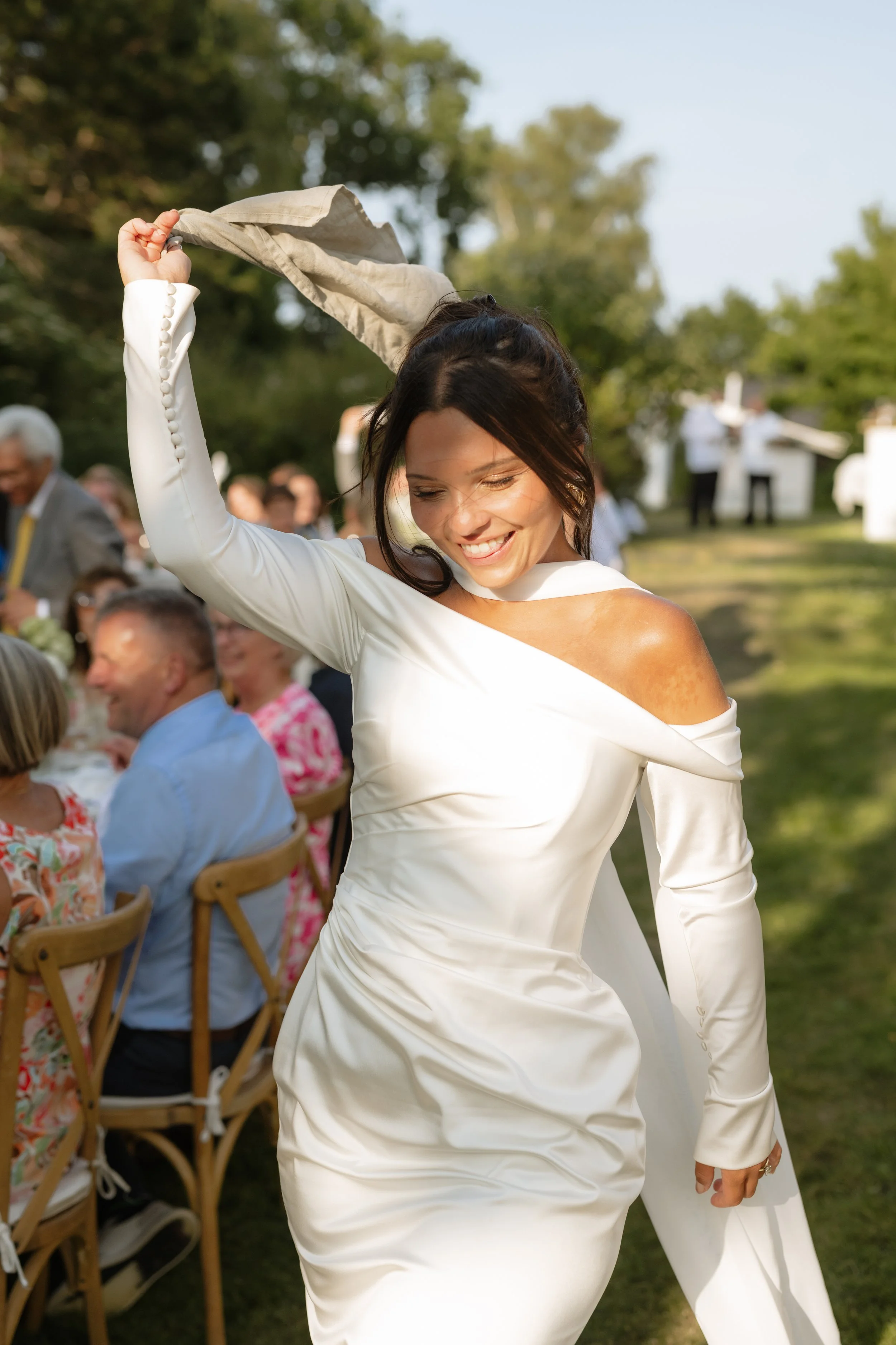 A woman in a white dress dance happily outdoors during a social gathering, with people sitting at tables and trees in the background.