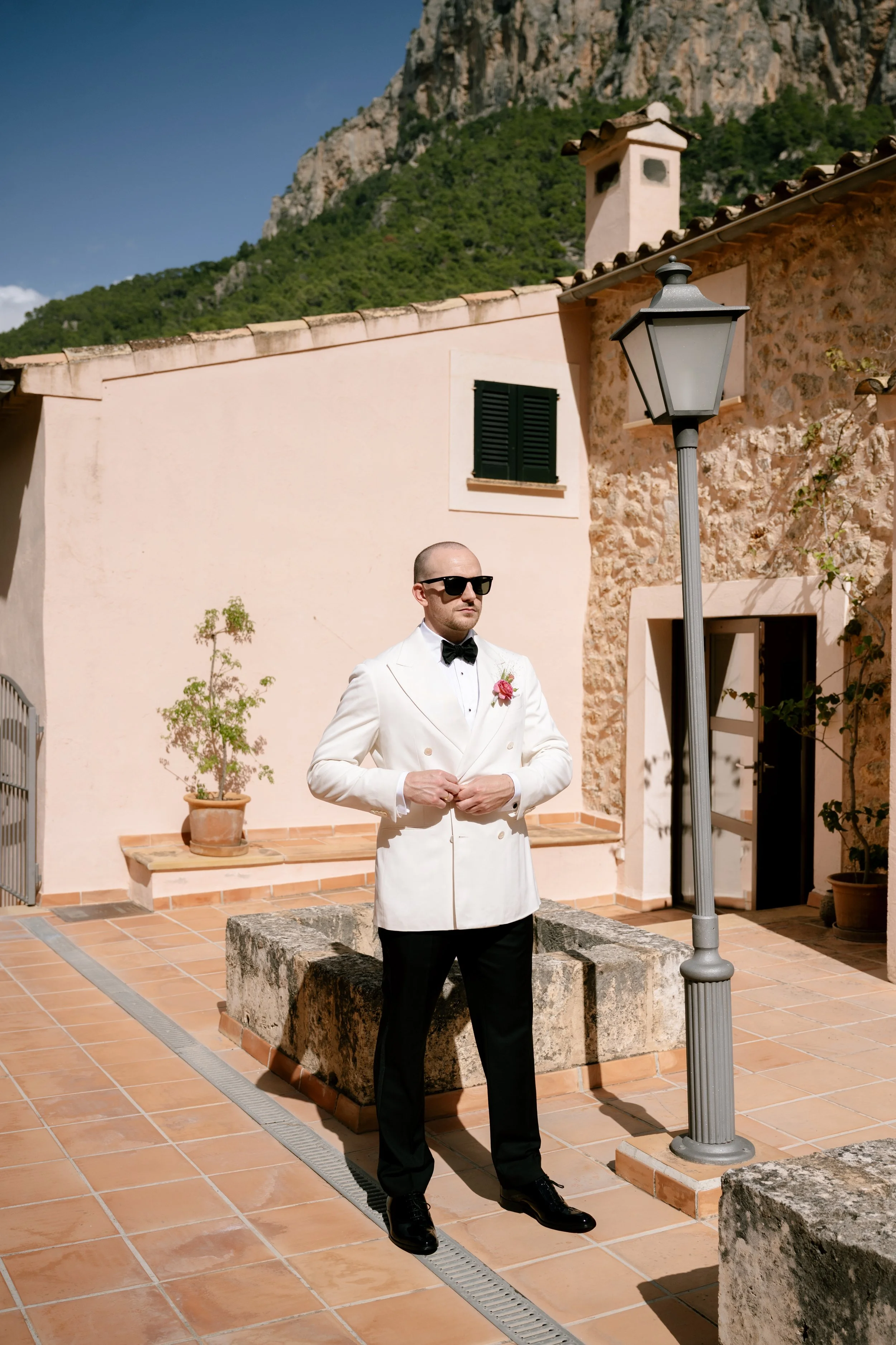 A man wearing a white tuxedo jacket, black bow tie, and sunglasses stands outdoors on a tiled patio with a mountain backdrop. He has a pink boutonniere on his lapel and is adjusting his cuffs next to a street lamp.