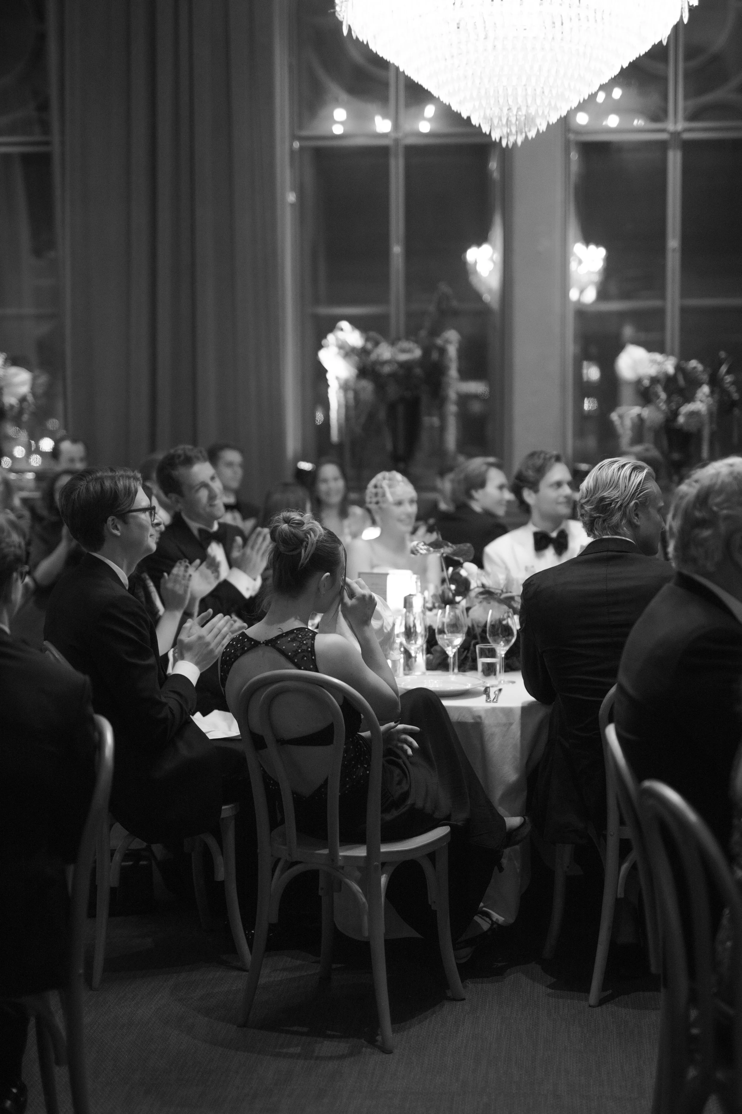 A black-and-white photo of an elegant reception or gala with people seated at round tables, dressed in formal attire, clapping and enjoying a speech or performance, in a decorated room with large windows, floral arrangements, and a chandelier.
