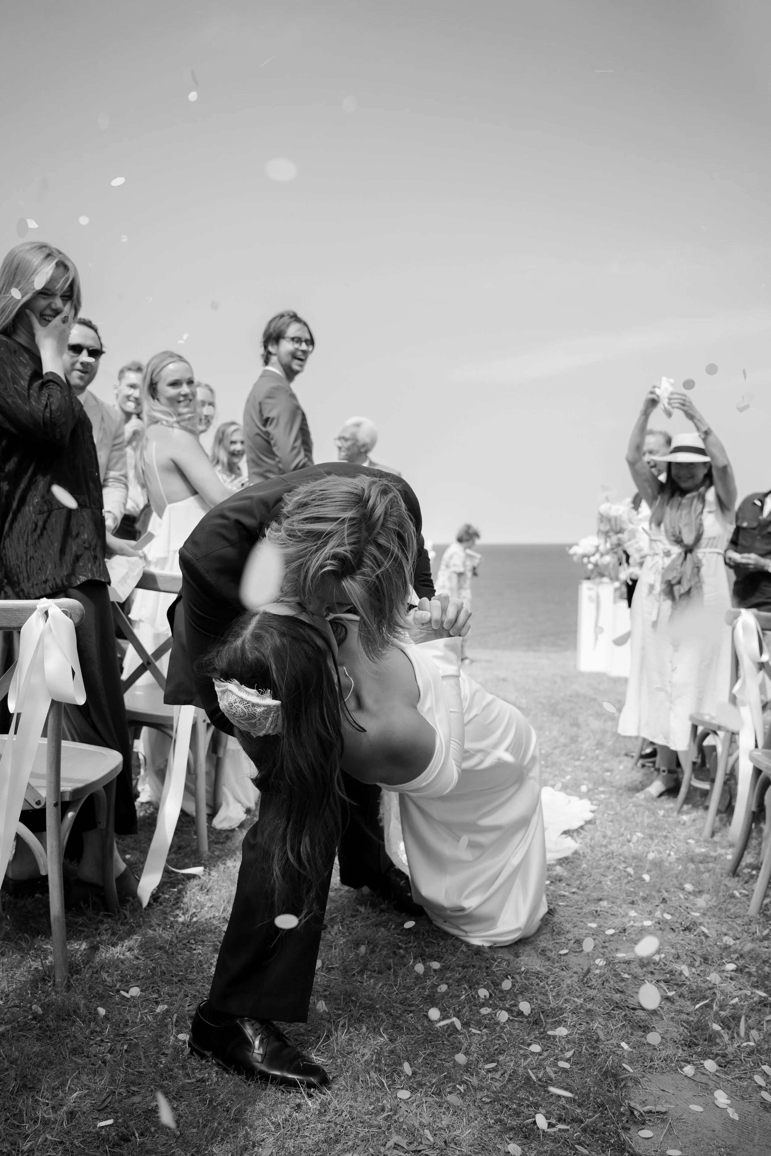 A wedding celebration on a beach with a bride and groom kissing, group of guests smiling and cheering, confetti falling.