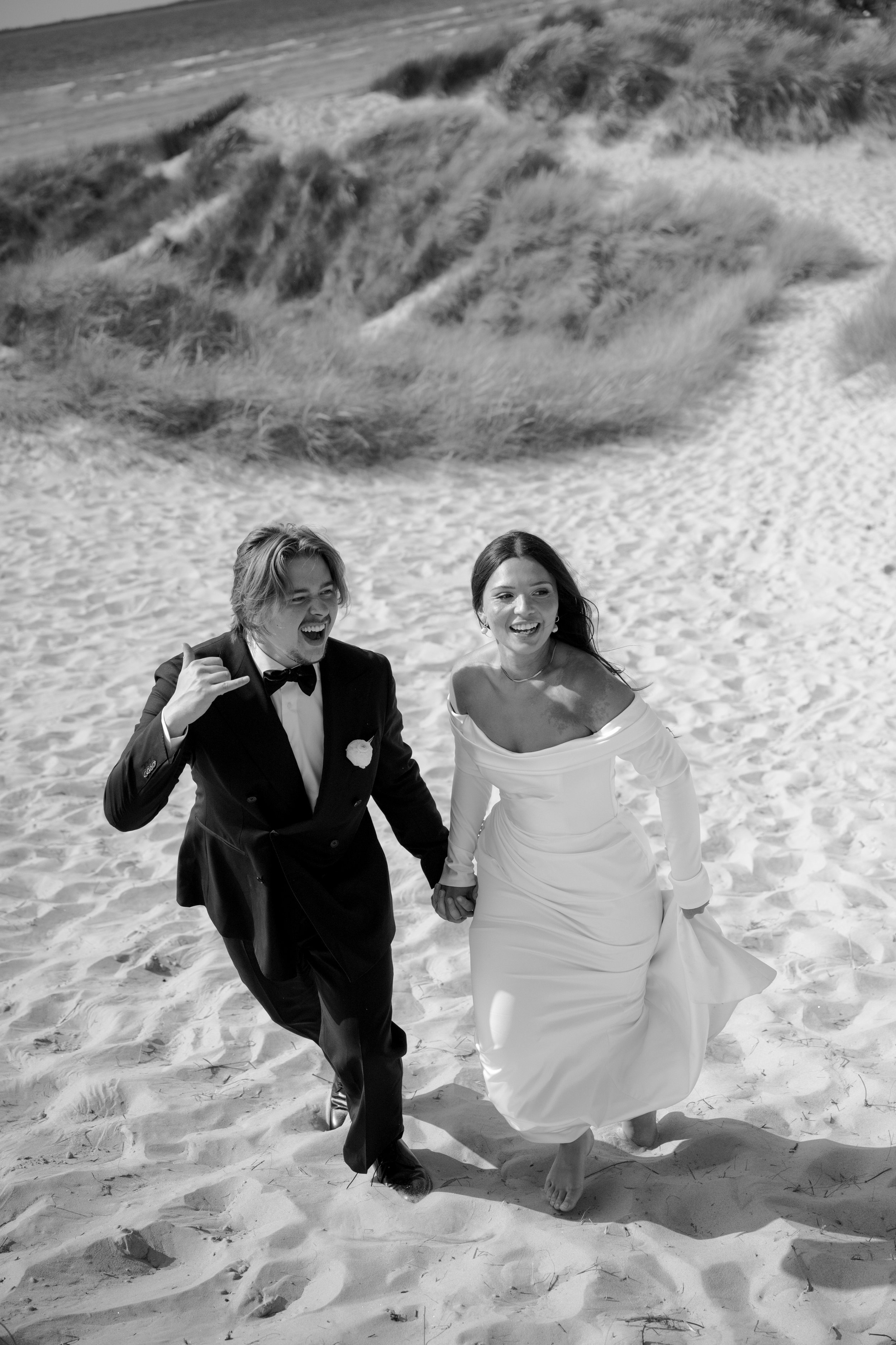 A newlywed couple in wedding attire running barefoot on a sandy beach, holding hands and smiling with the dunes and grass in the background, in black and white.