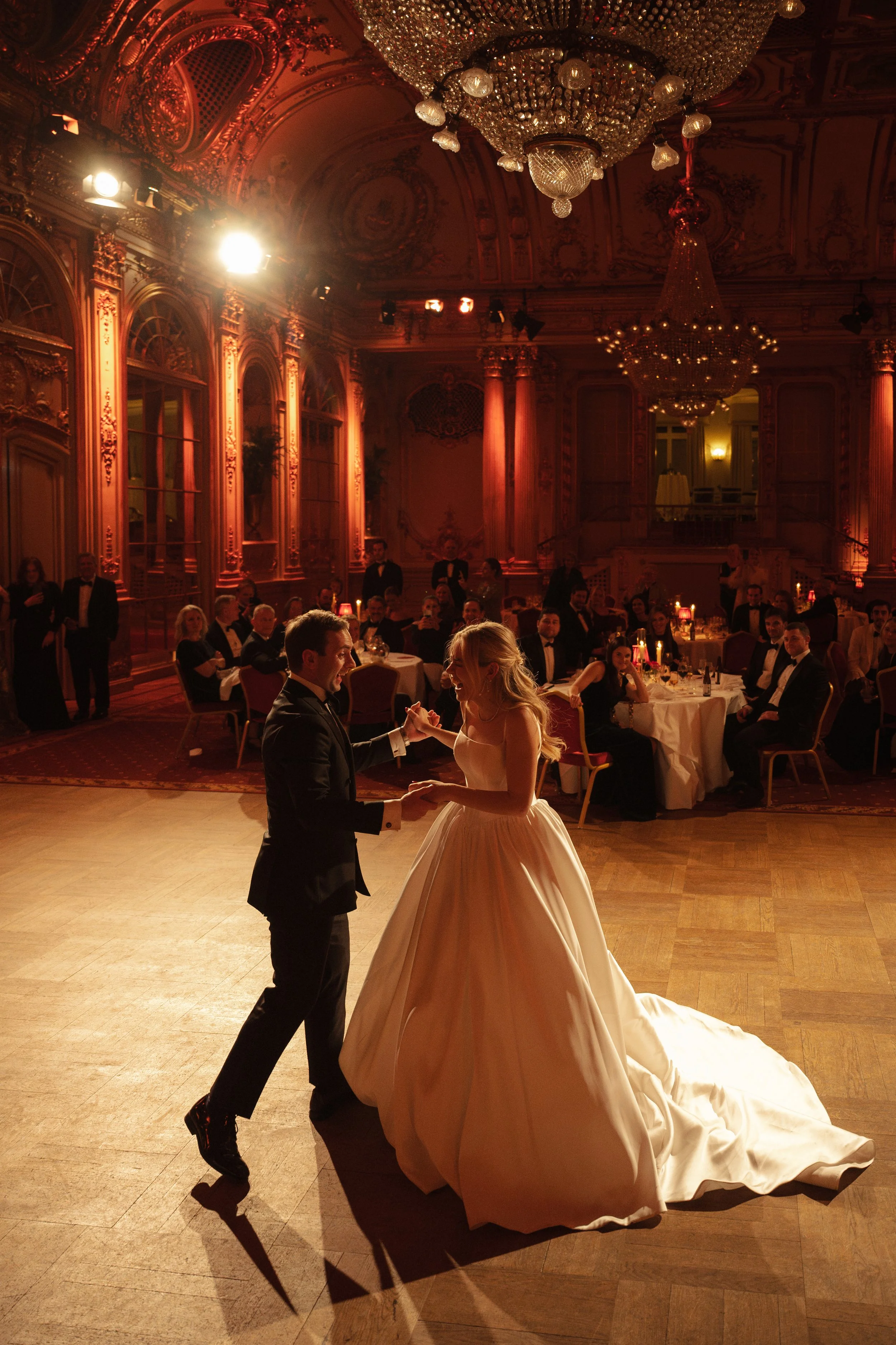 A bride and groom dancing at their wedding reception in an elegant, ornate ballroom with chandeliers and guests seated at decorated tables.