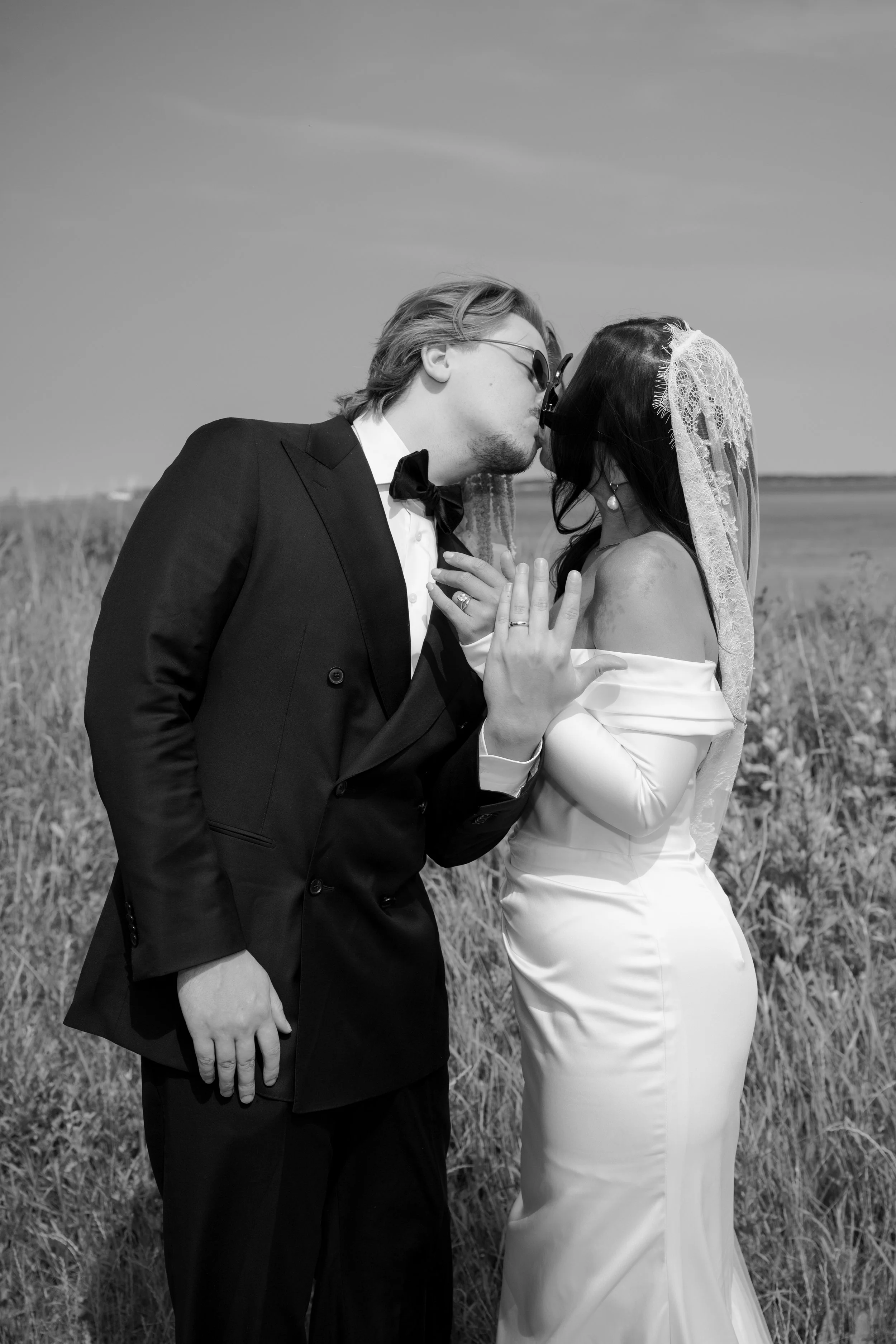 A black-and-white photo of a couple in wedding attire kissing outdoors in a field with a body of water in the background.