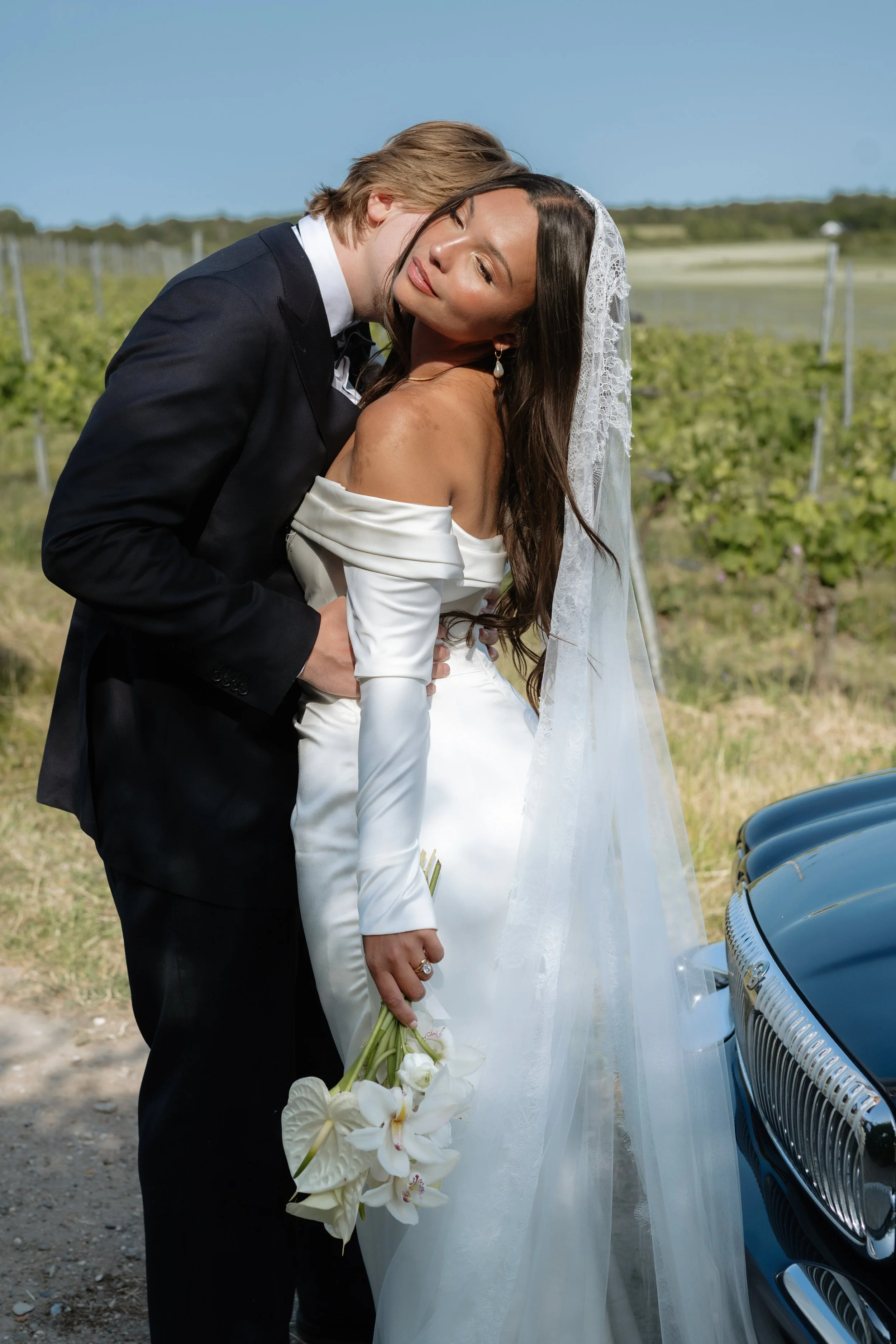 A newlywed couple sharing a kiss outdoors, with a bride in a white gown and veil holding a bouquet of white flowers, and a groom in a black tuxedo, near a vintage car, in a scenic rural setting with vineyard rows in the background.
