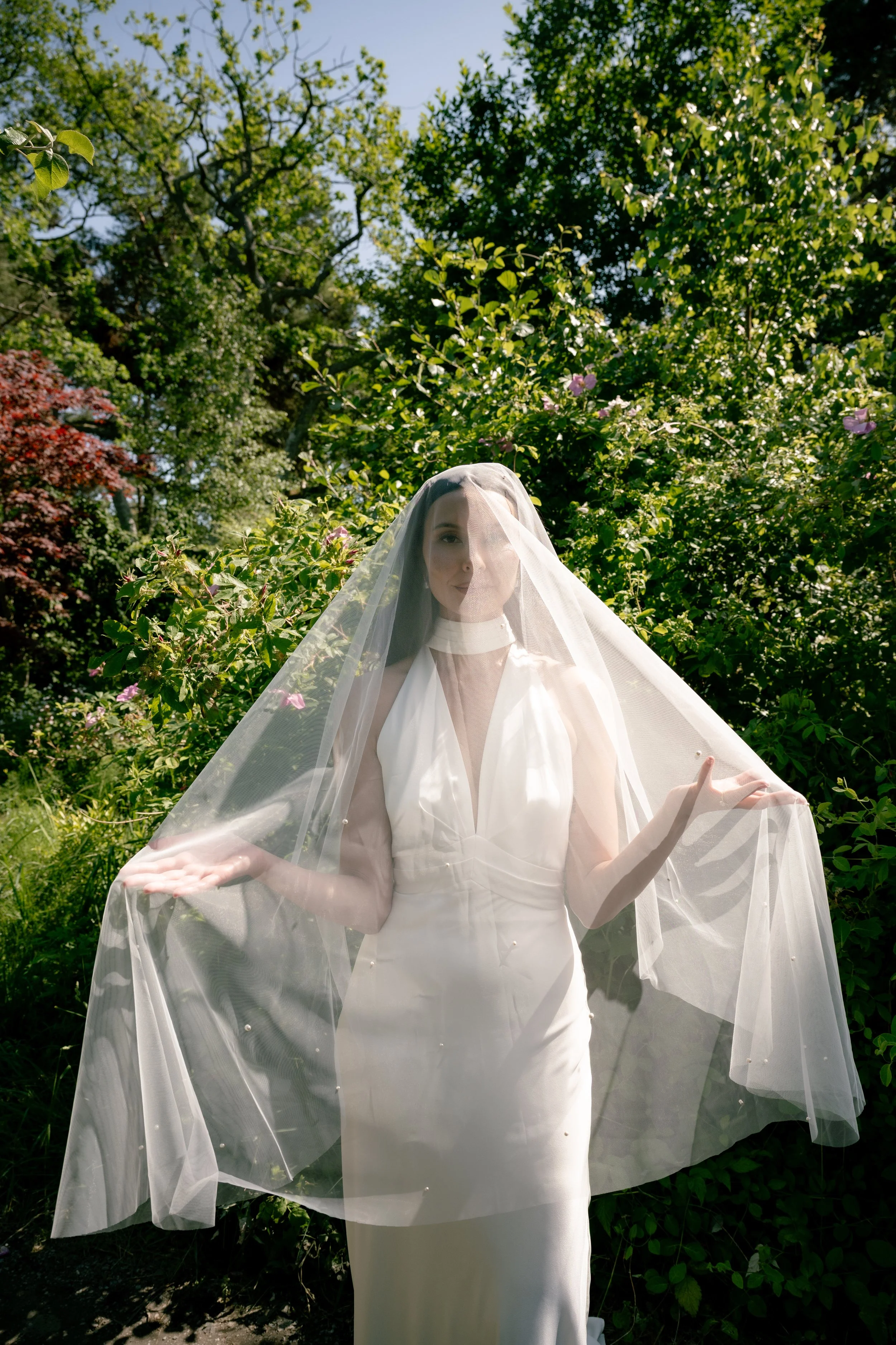 A woman dressed in a white evening gown with a sheer veil over her head and shoulders, standing amidst green bushes and trees on a sunny day.