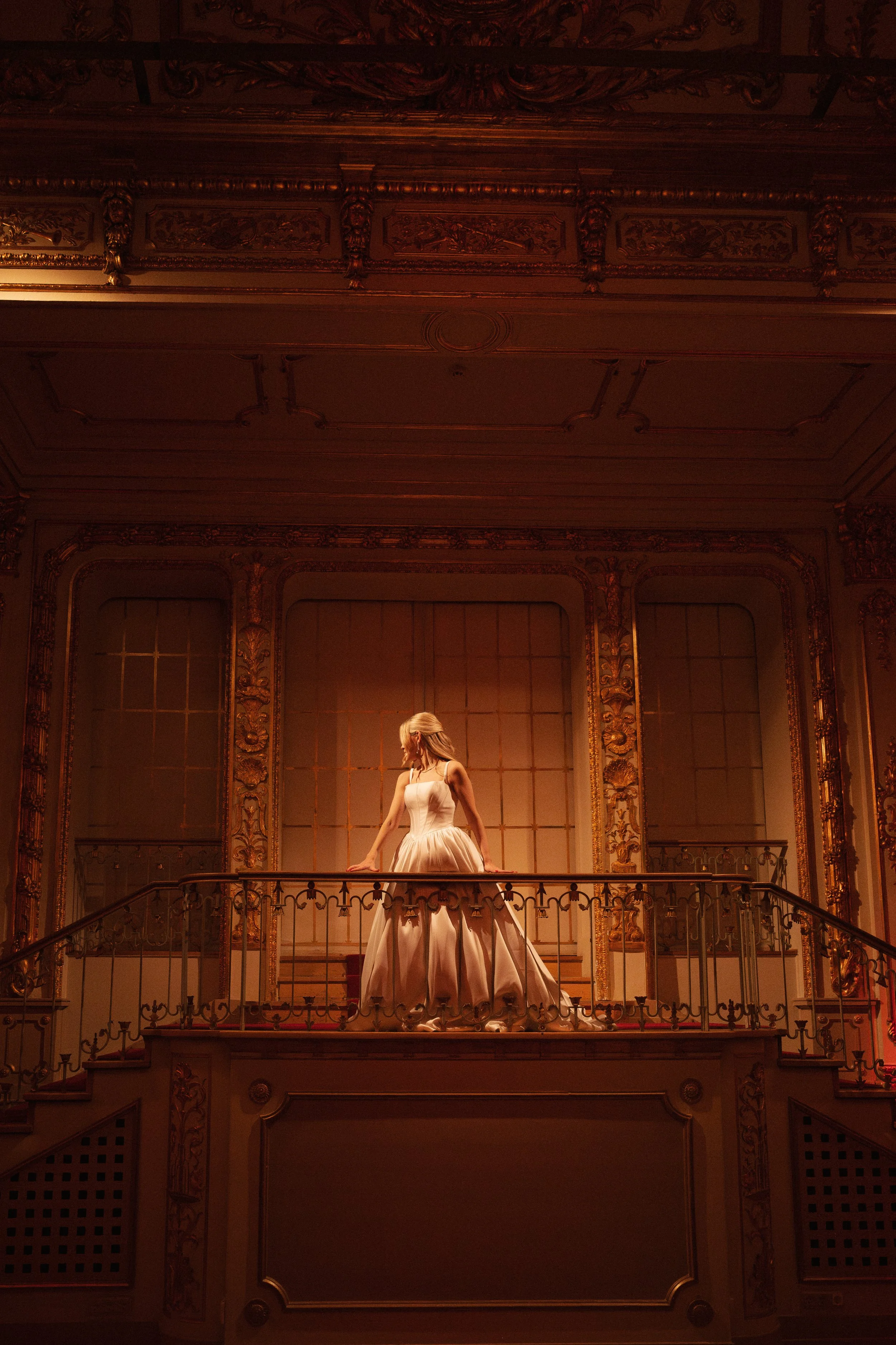 A woman in an elegant, strapless gown standing on a balcony in a richly decorated room with gold accents and ornate carvings.