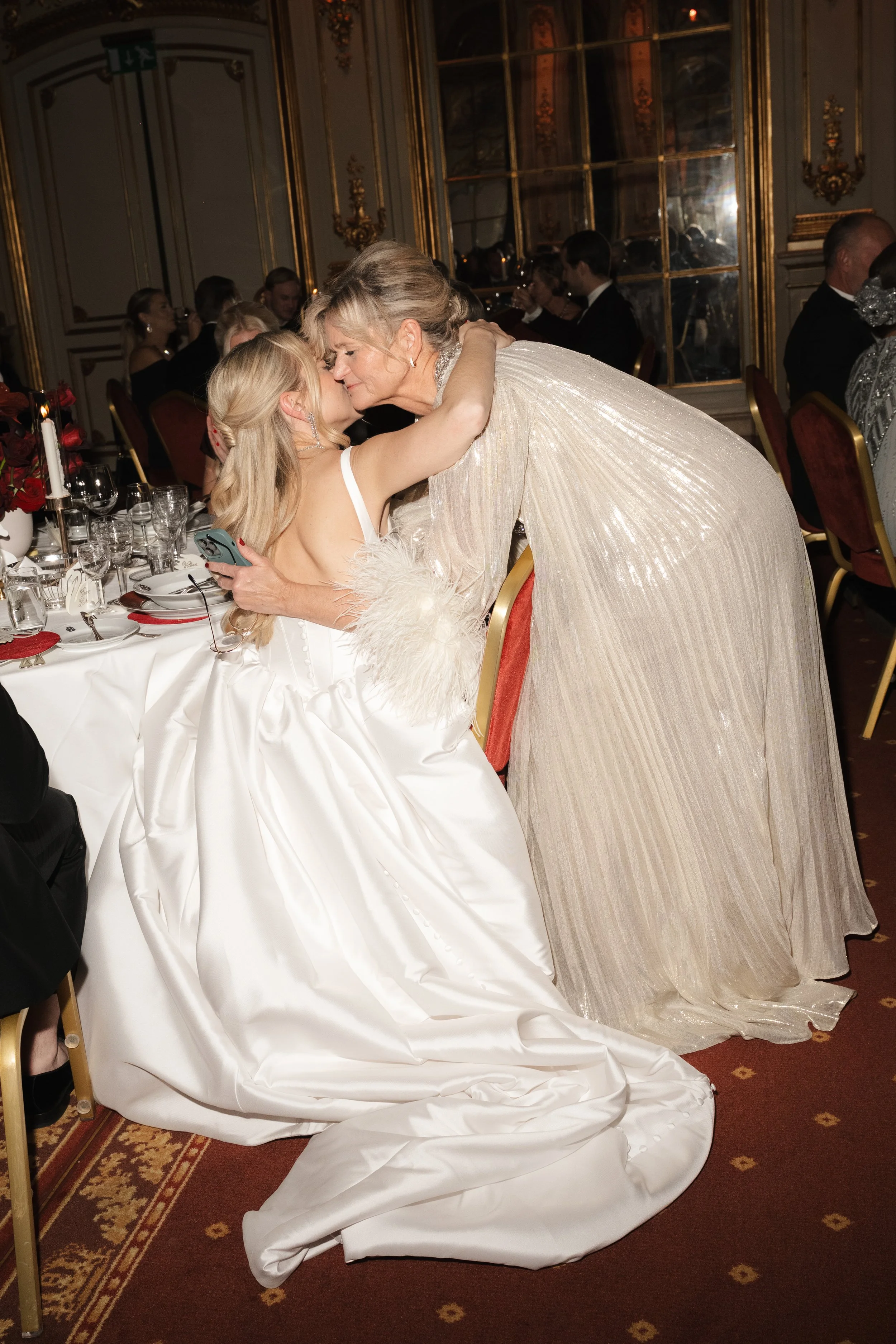 A young girl in a white dress and an older woman in a shiny, beige gown share a warm embrace at a formal banquet, surrounded by guests at decorated tables in an elegant, ornate room.