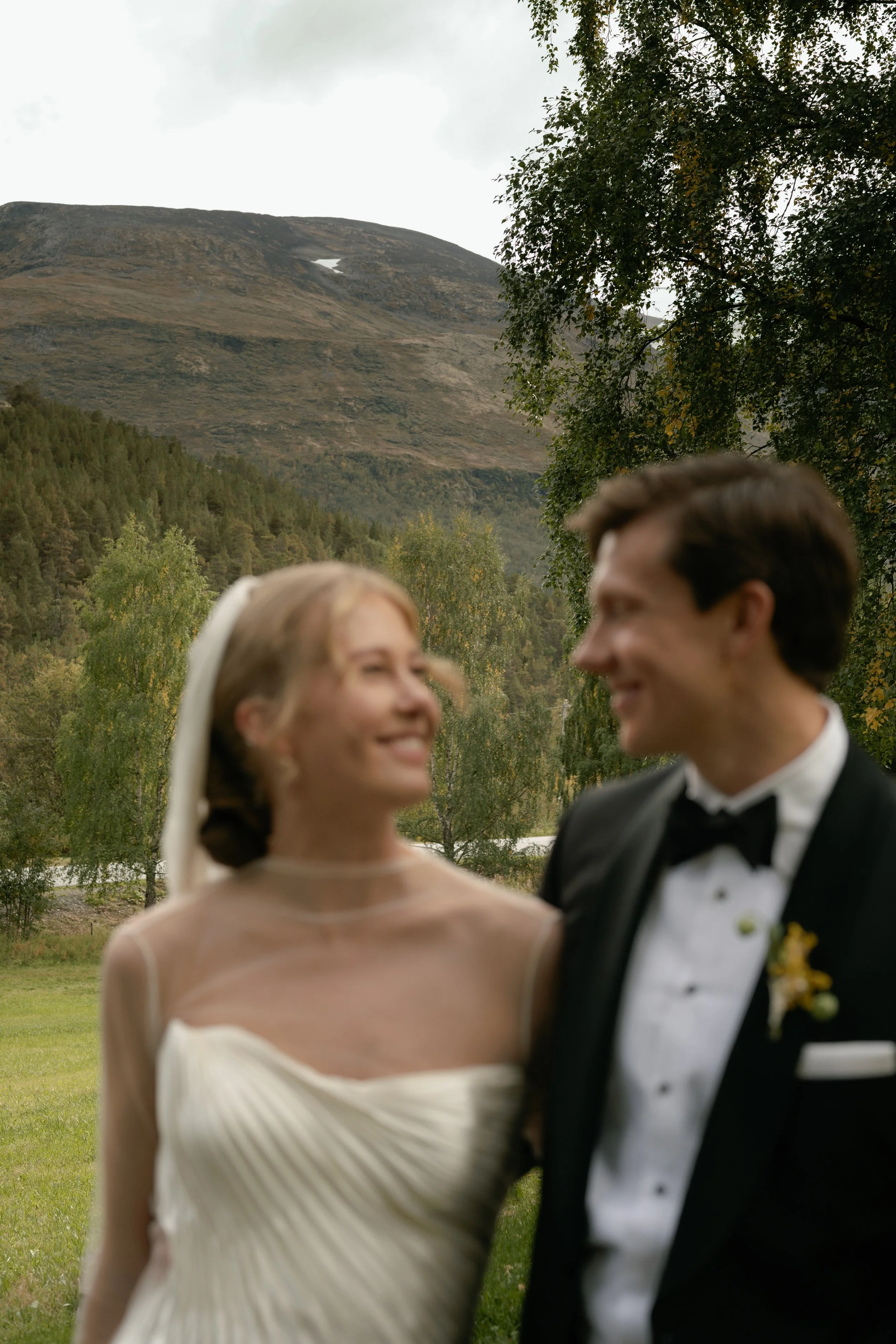 A couple dressed in wedding attire standing outdoors in front of mountains and greenery, smiling at each other.
