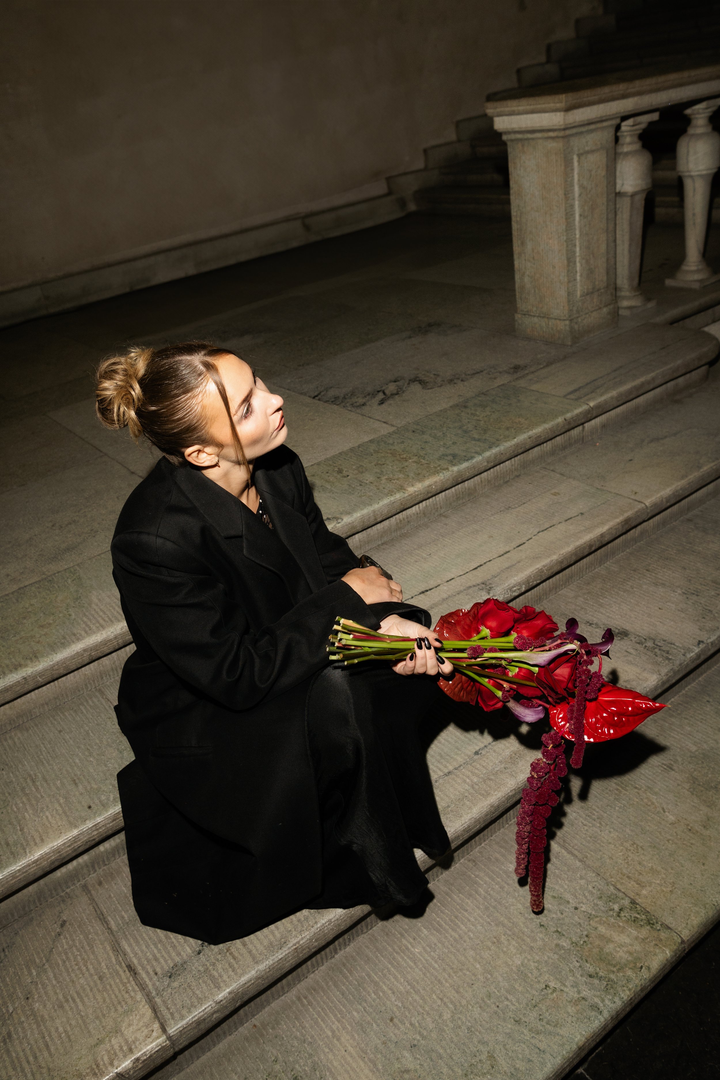 A woman sitting on stone stairs holding a bouquet of red and purple flowers, wearing a black coat, with her hair in a bun, in a dimly lit area.