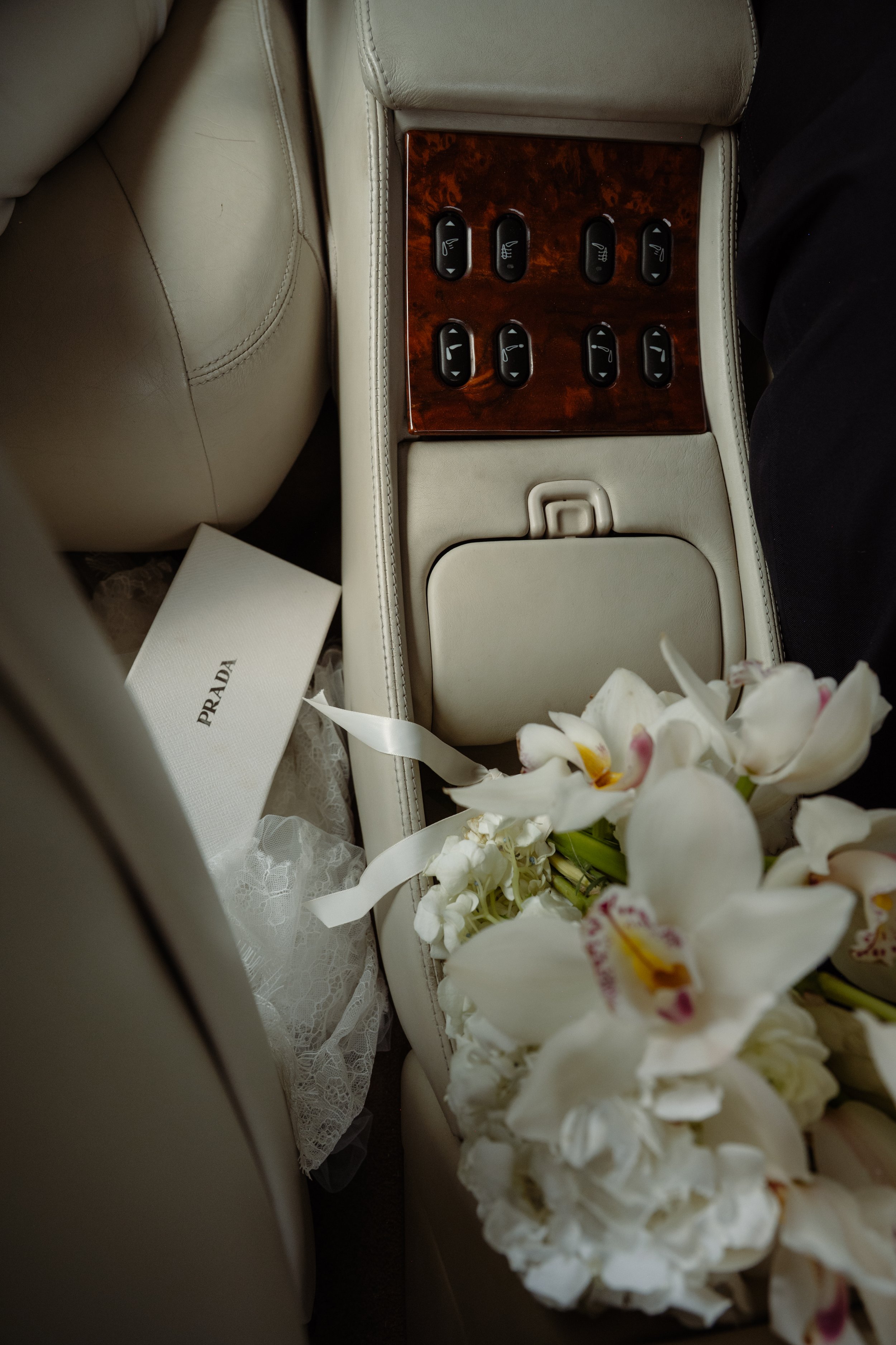 The back seat of a car with beige leather interior, showing a wooden panel with multiple black buttons, a beige storage compartment, a white prada box, a sheet of lace fabric, and a bouquet of white orchids and flowers.