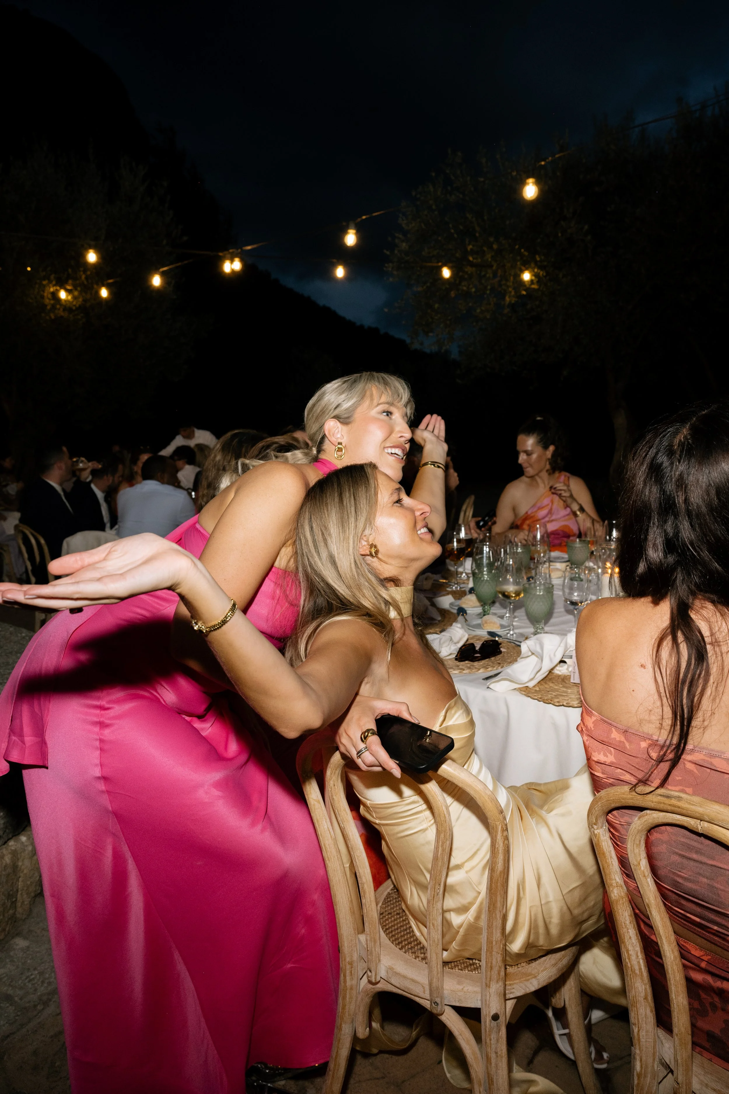 Women celebrating at an outdoor evening gathering with string lights and a mountain backdrop.