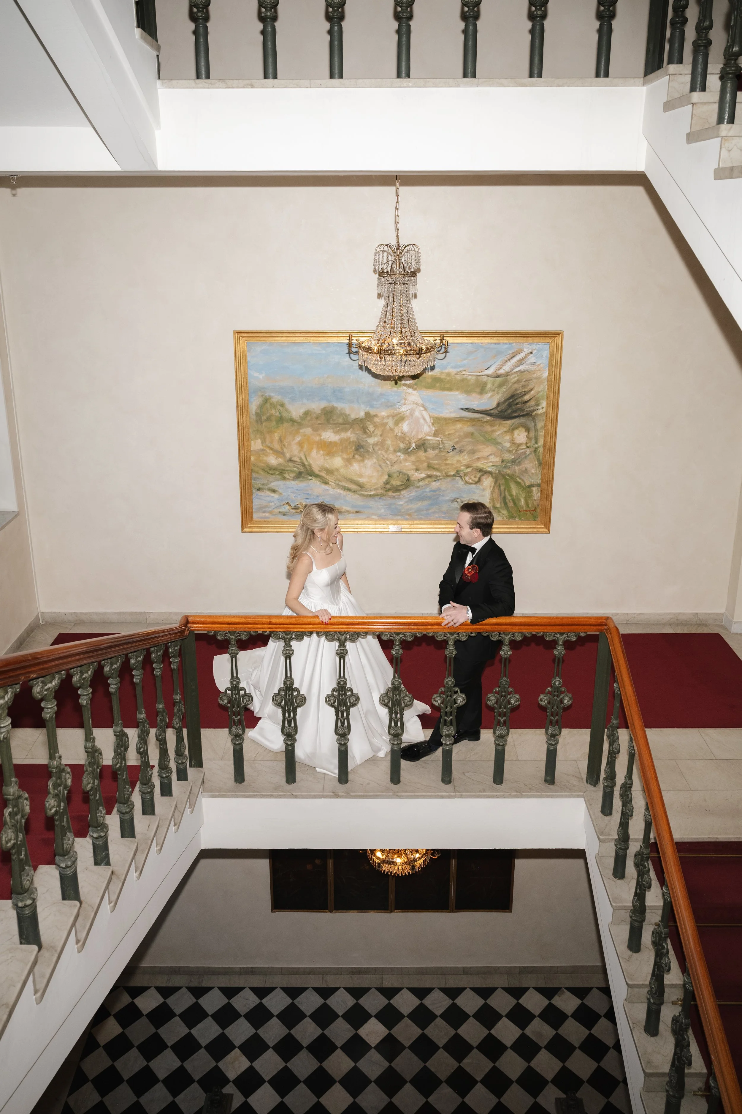 A bride and groom in wedding attire talking on a staircase landing, with a chandelier above and a landscape painting behind them.