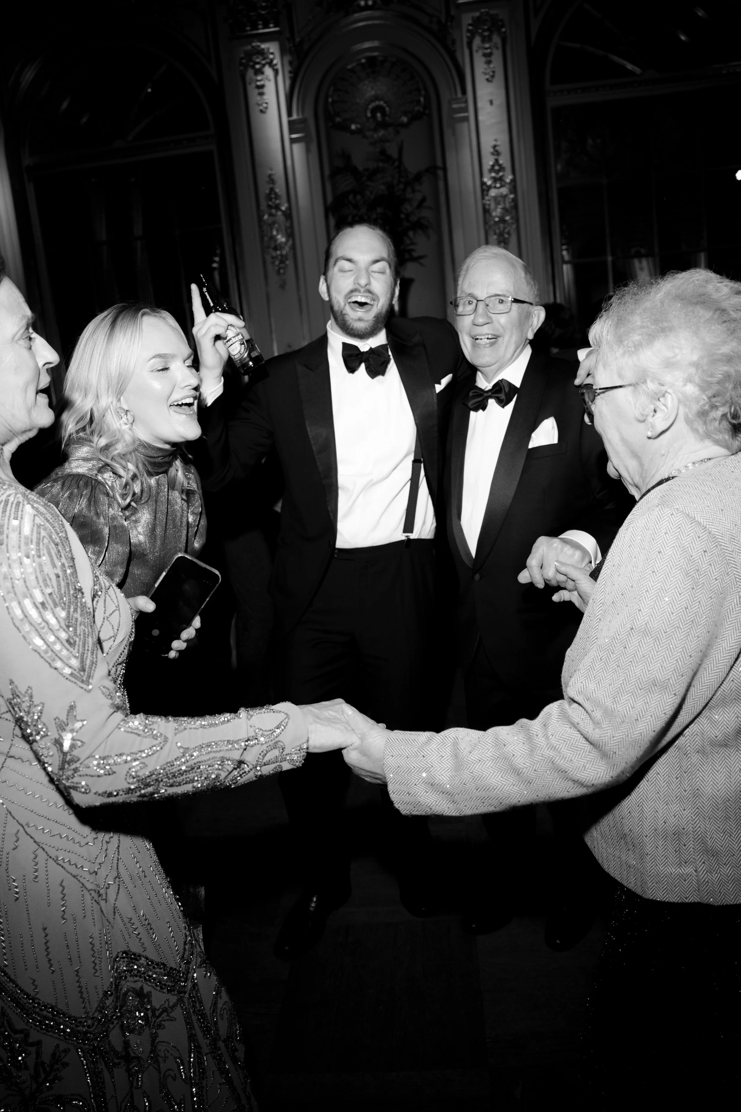 People dancing and celebrating at a formal event in an elegant room, with men in tuxedos and women in dresses, smiling and engaging in lively interaction.
