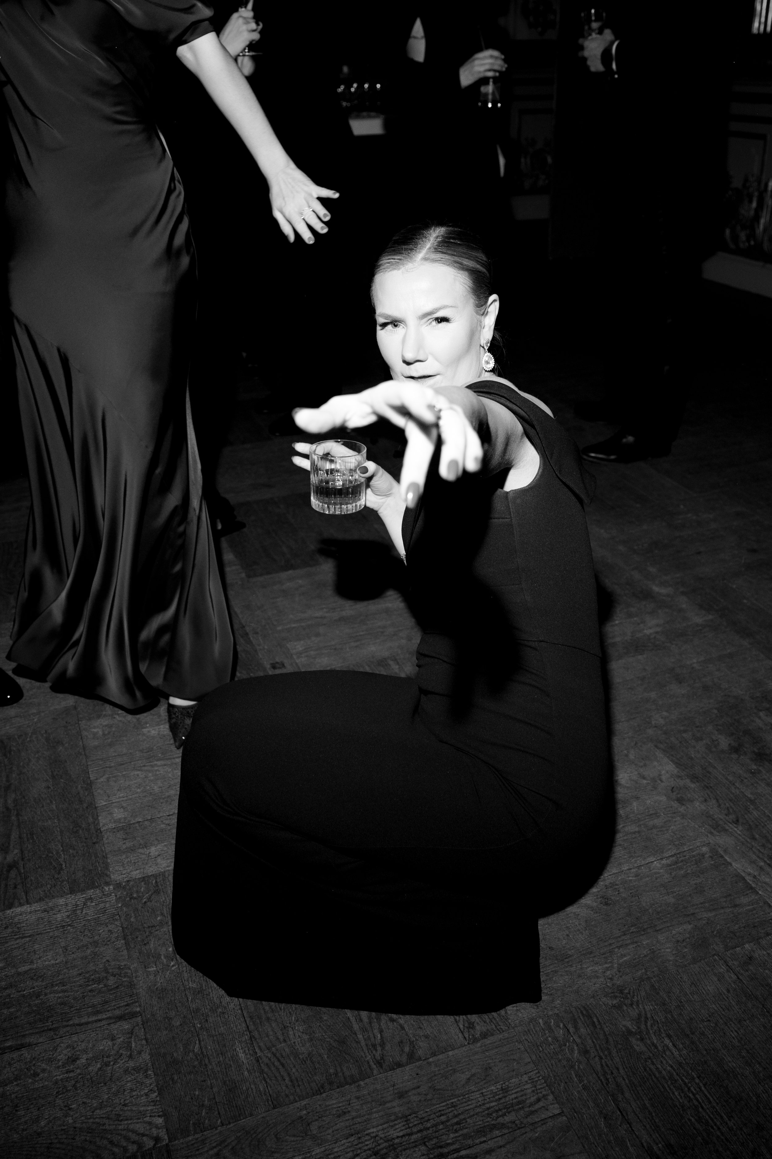 A woman in a black dress kneeling on the dance floor, holding a glass of drink in one hand, and pointing toward the camera with a playful expression at a party or celebration.