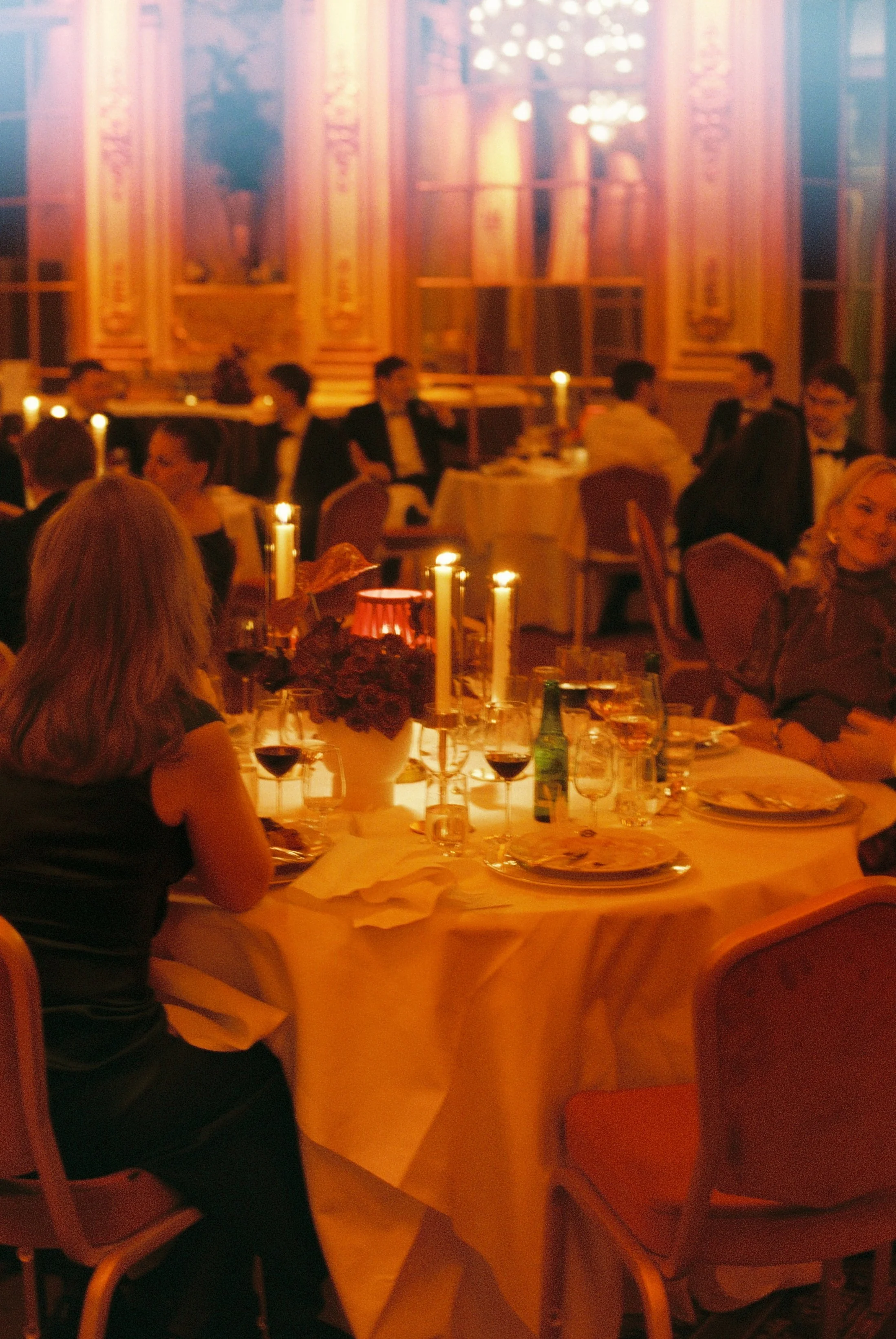 People dining at a formal event with candles and elegant table settings in a luxurious, ornate room with gold accents and chandelier lighting.