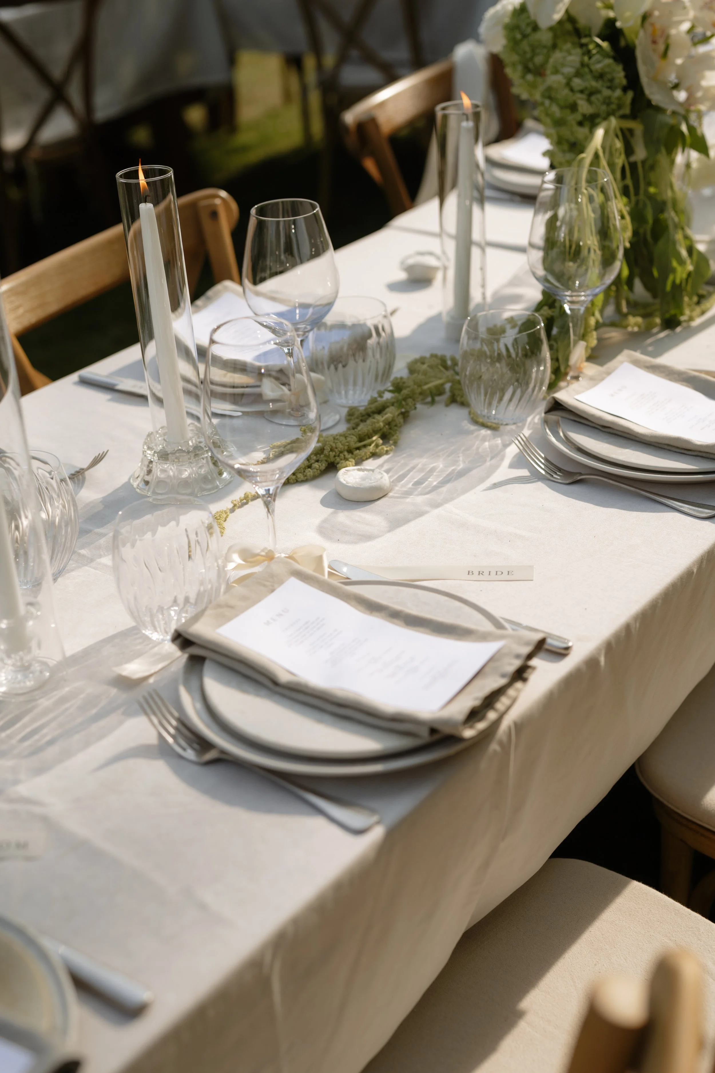 Elegant outdoor wedding table setting with white tablecloth, wine glasses, candles, floral arrangements, and place cards.