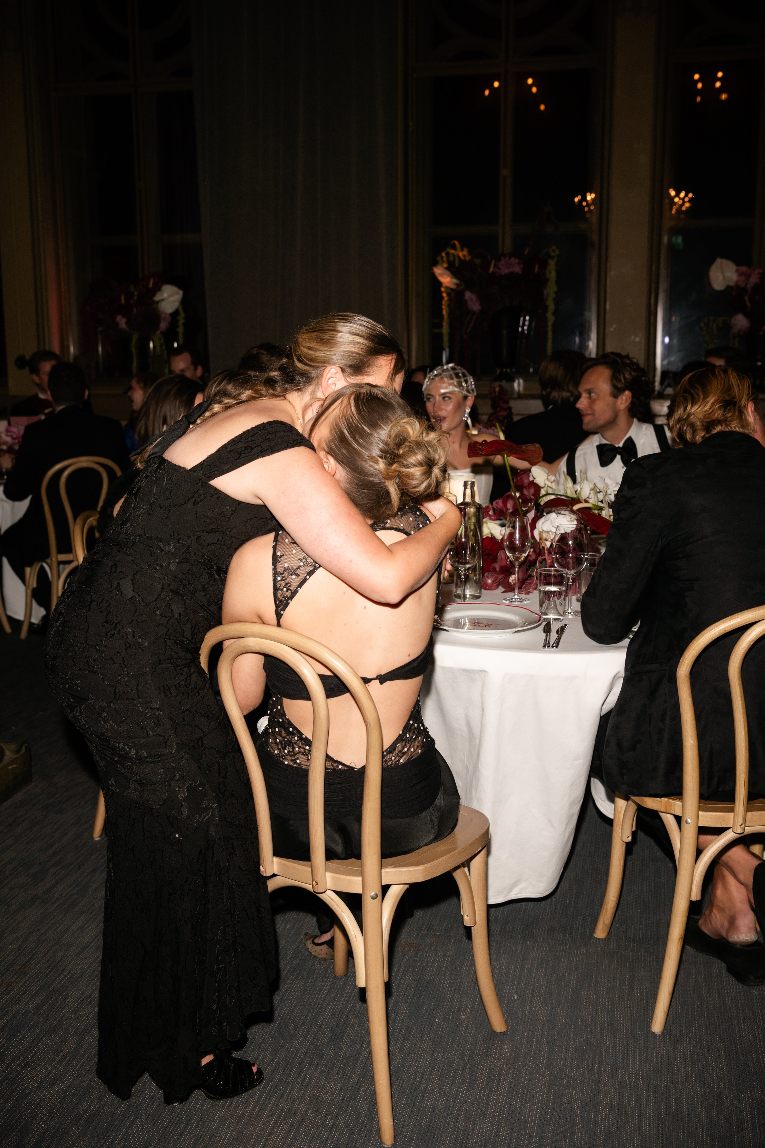 People at a formal event, including a woman embracing another woman in black lace eveningwear, seated at a table with elegant floral arrangements and dinnerware.