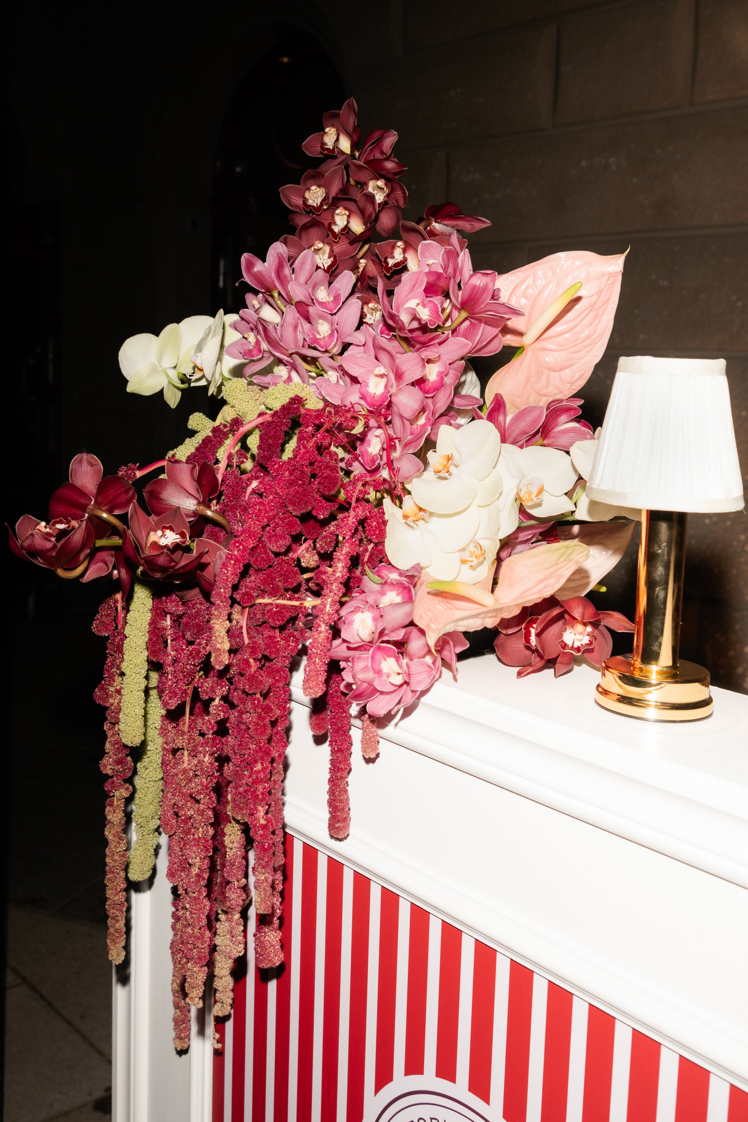 A floral arrangement featuring pink, white, and burgundy orchids, with an elegant gold and white lamp on a white surface, against a dark wall with red and white striped decor.