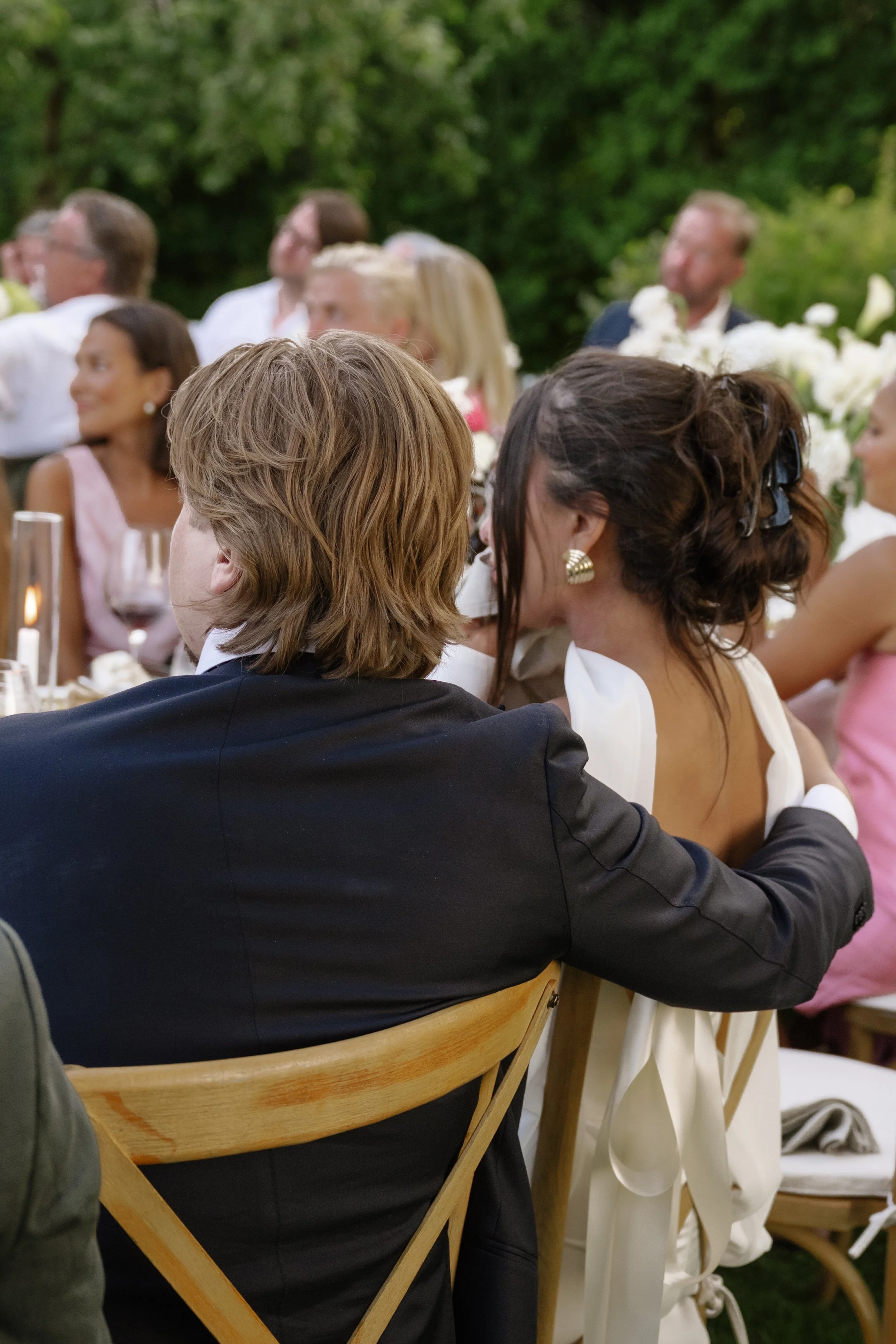 People sitting at a wedding reception outdoors, with a man in a dark suit and a woman in a white dress in the foreground.