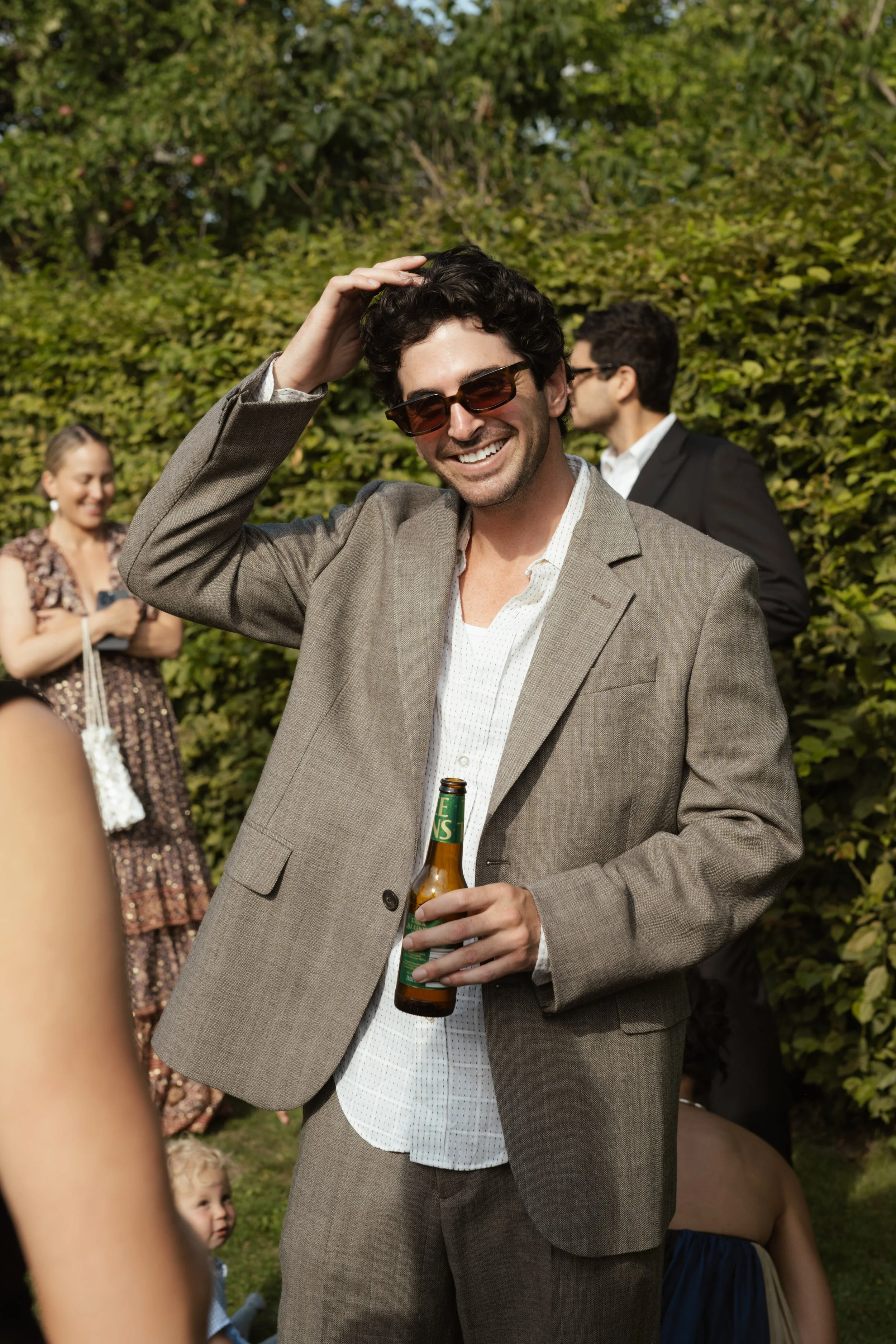 A man wearing sunglasses and a gray suit is smiling, holding a beer bottle, and touching his hair at an outdoor gathering.