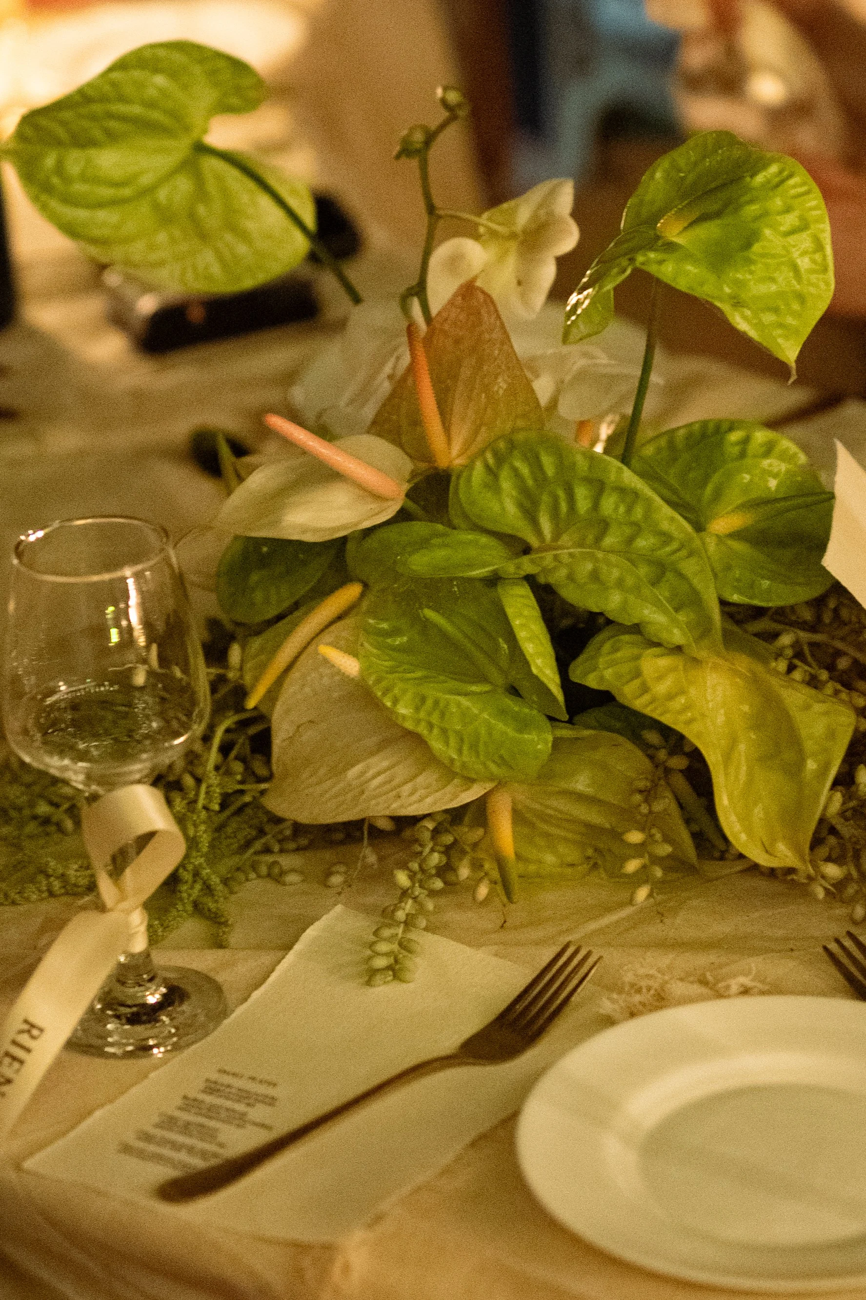 A dining table setting with a centerpiece of green and brown leafy plants, a white flower, an empty wine glass with a tag, a fork, a white plate, and a printed menu.