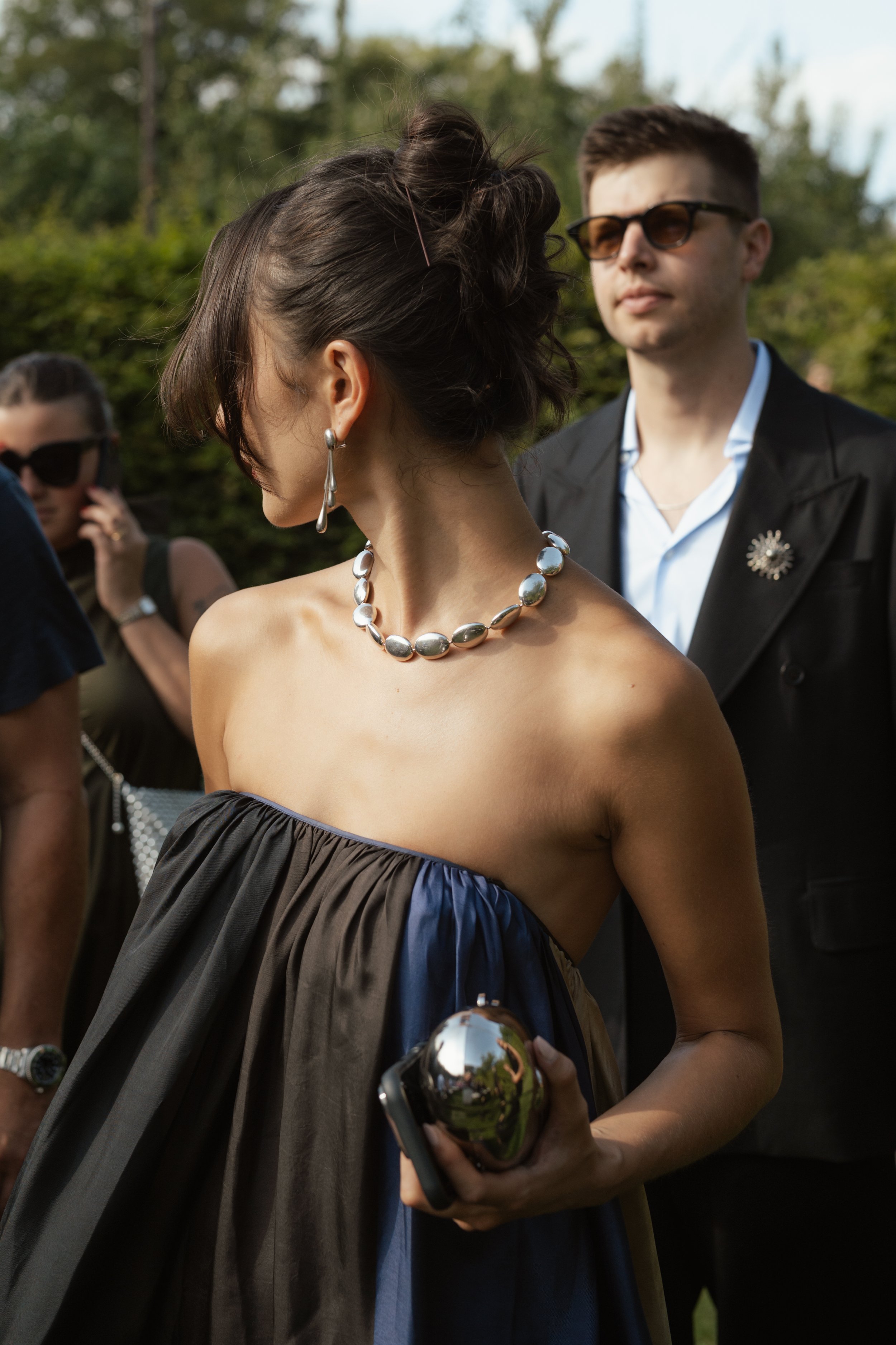 A woman in an off-shoulder black dress with a silver necklace and earrings, holding a silver clutch, standing outdoors with people in the background, and a man in sunglasses standing behind her.