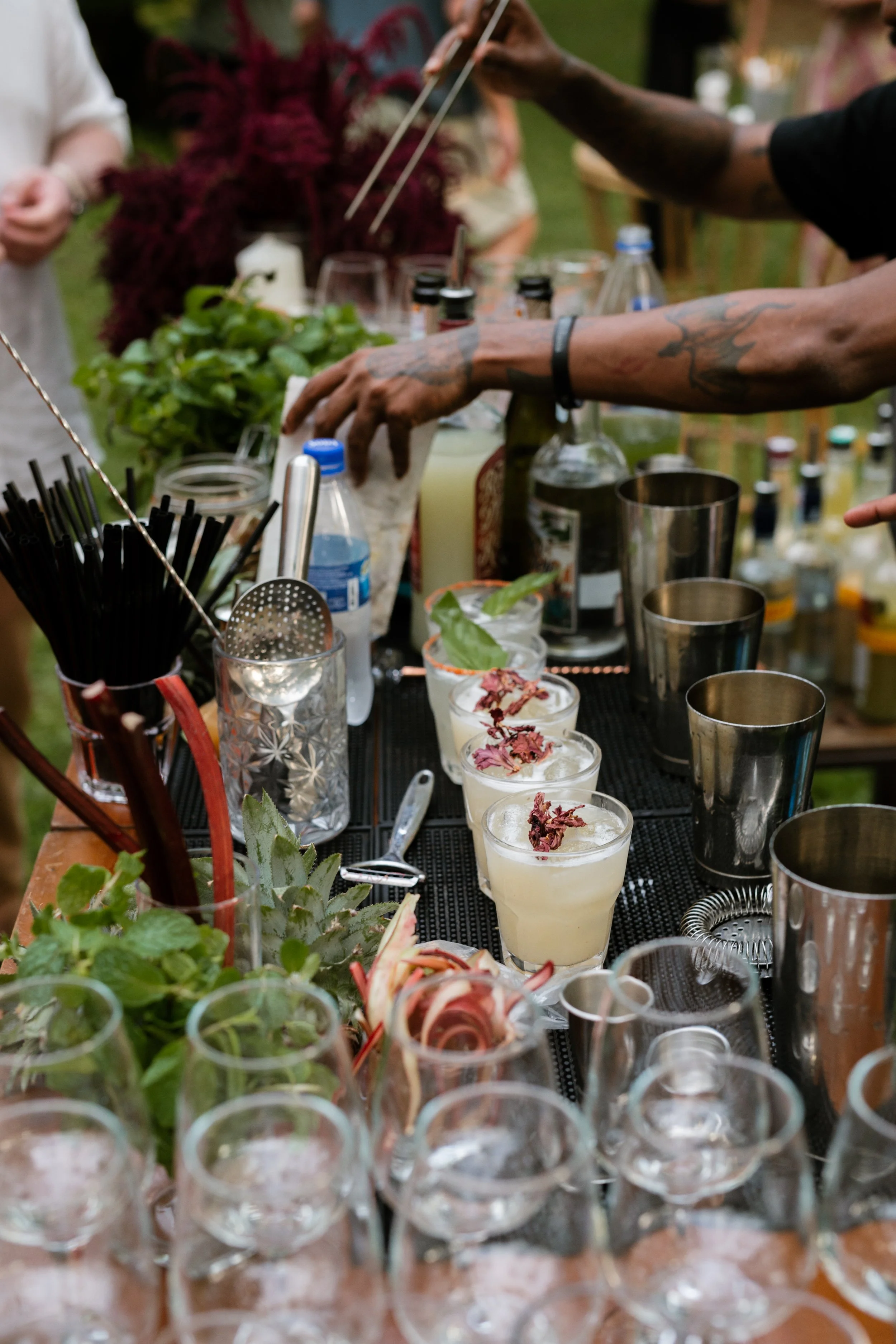 A bartending setup with various liquor bottles, glasses, garnishes, and mixing tools on an outdoor bar table, with people in the background.