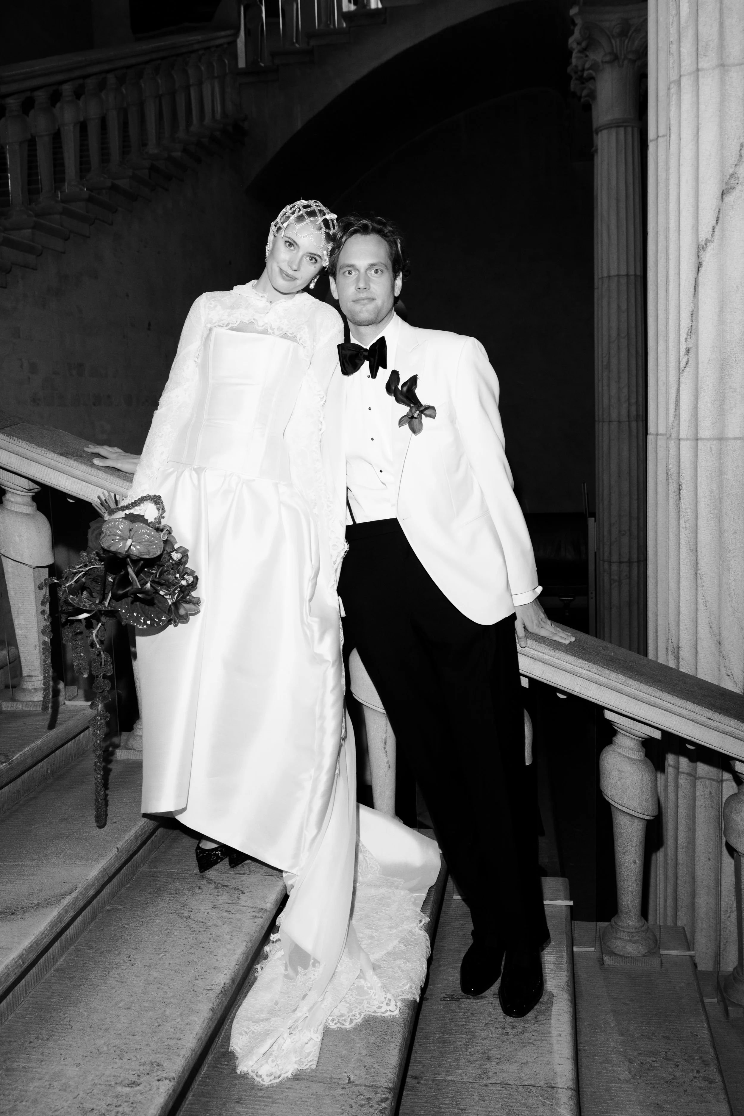 Black and white photo of a bride and groom on a staircase, with the bride in a wedding gown holding a bouquet, and the groom in a tuxedo with a bow tie, near a decorative railing and classical columns.