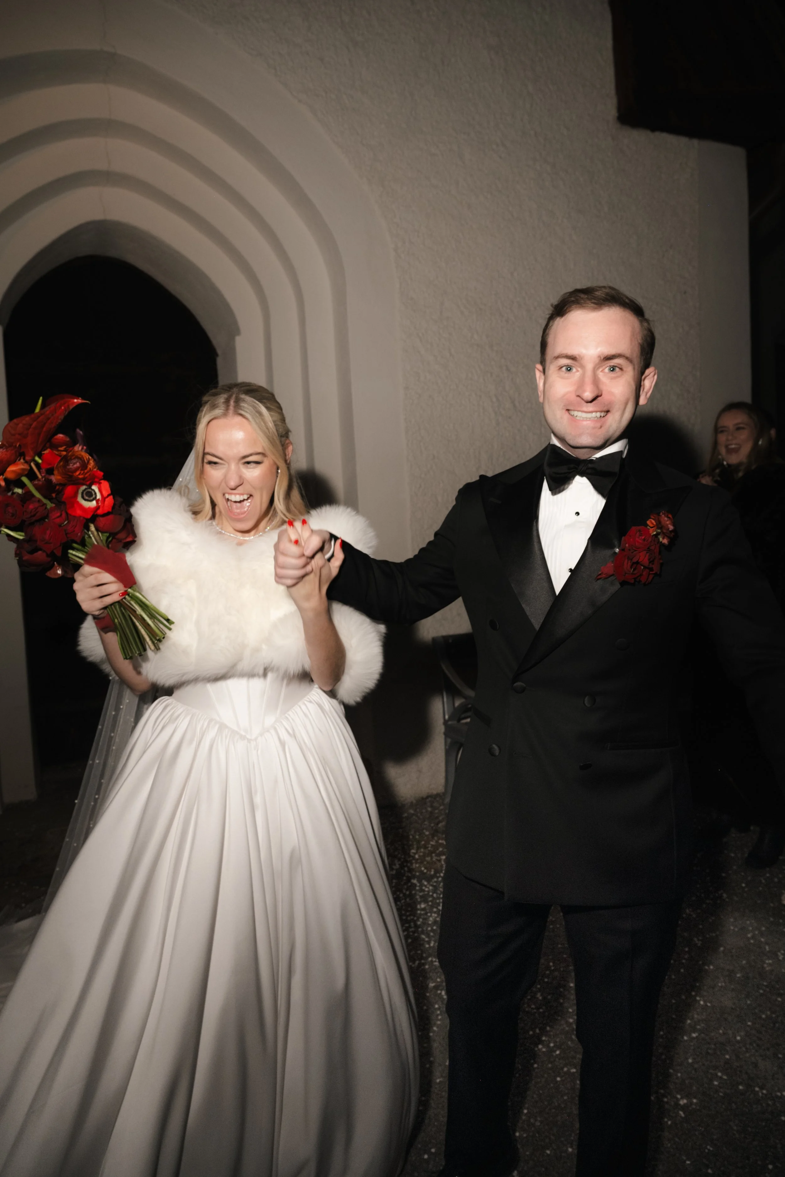 A newlywed couple holding hands and smiling at their wedding reception, with the bride in a white gown and holding a bouquet, and the groom in a tuxedo with a red boutonniere.