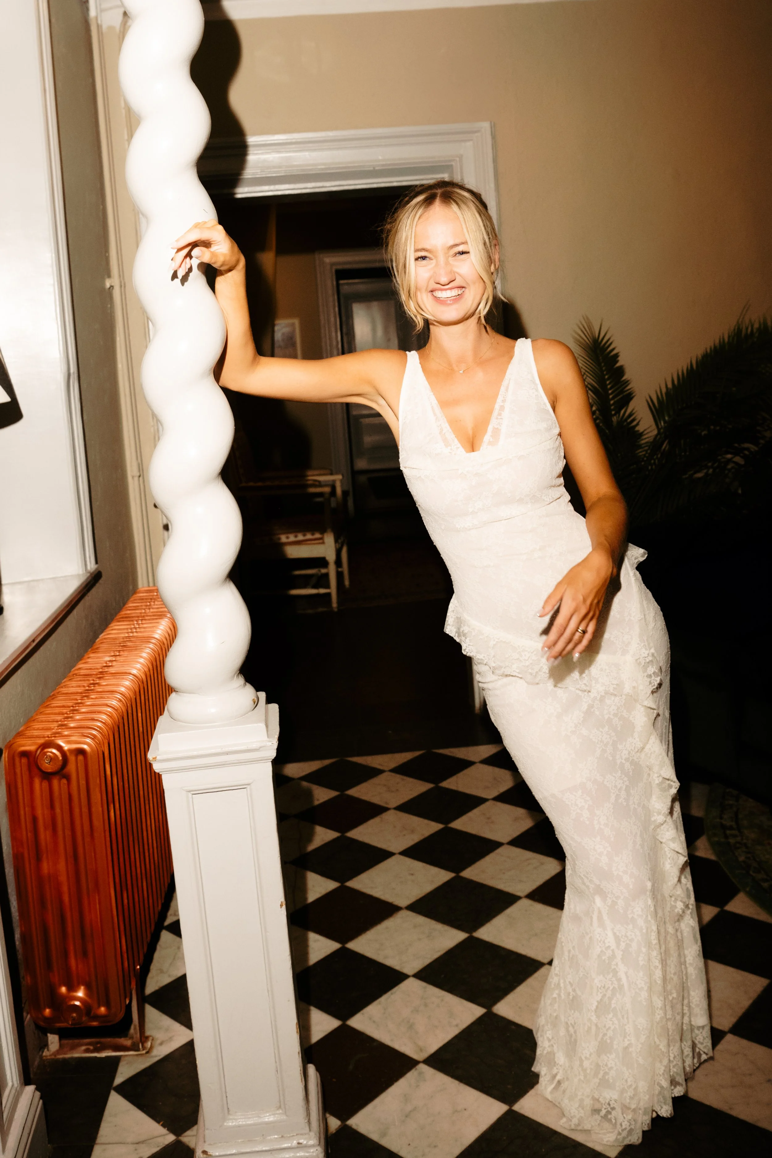 A woman in a white lace dress smiling and posing indoors, holding onto a decorative white twisted column with her right hand.