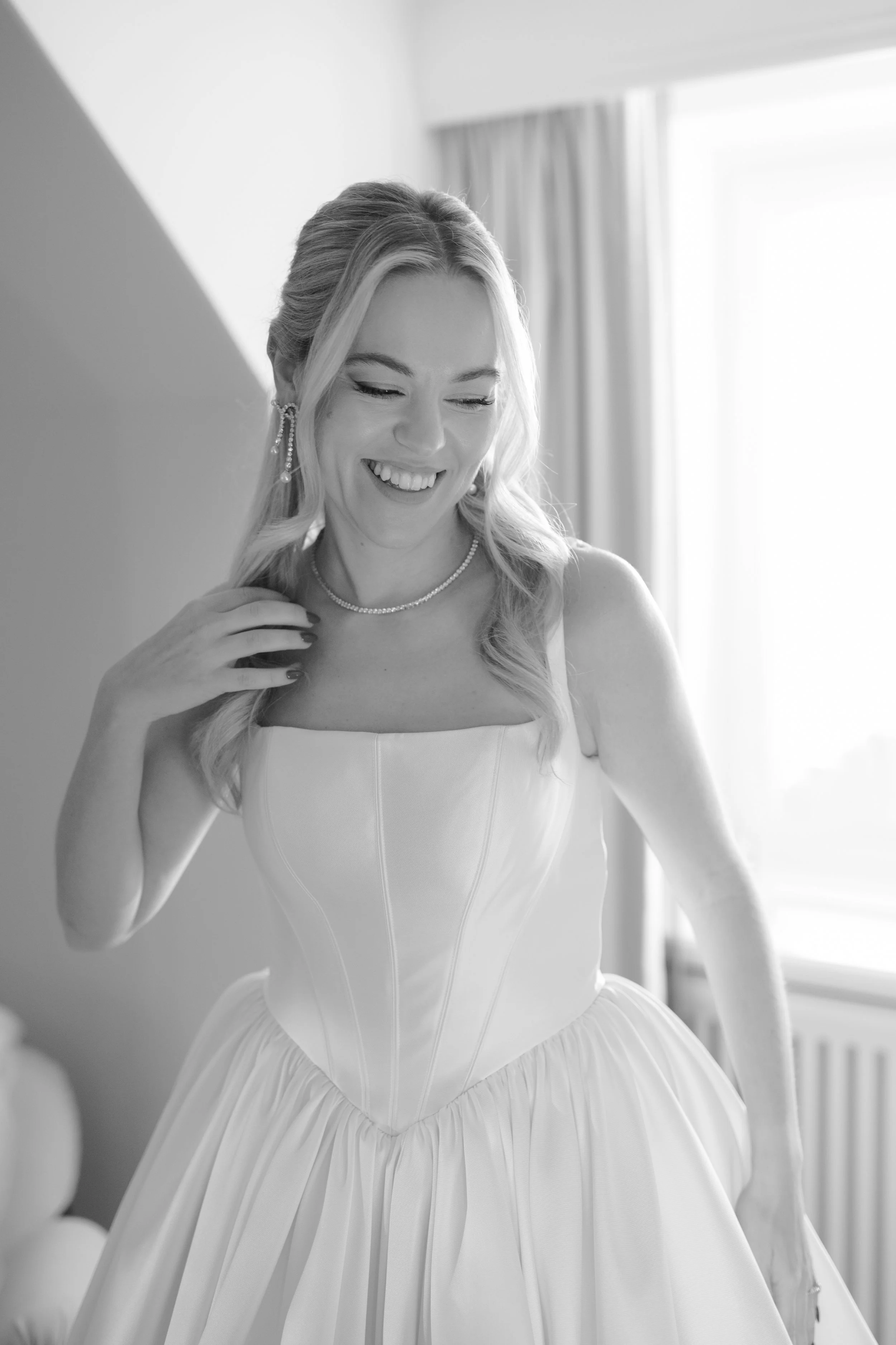 Black and white photo of a woman in a wedding dress smiling, with hand near her face, in a room with sunlight and curtains.