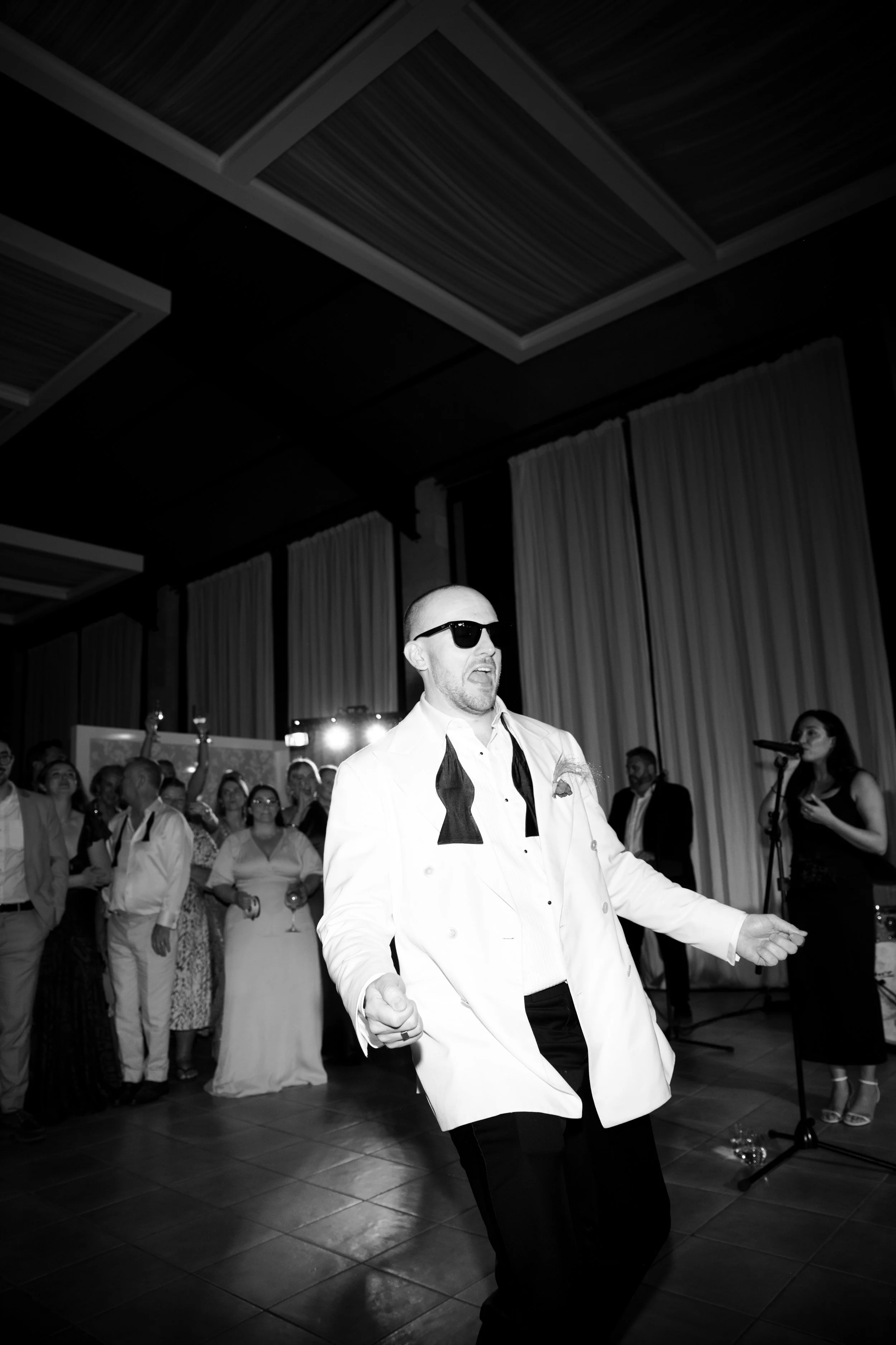 A man in sunglasses and a white tuxedo with a black bow tie dancing at a party or wedding reception with guests in the background.