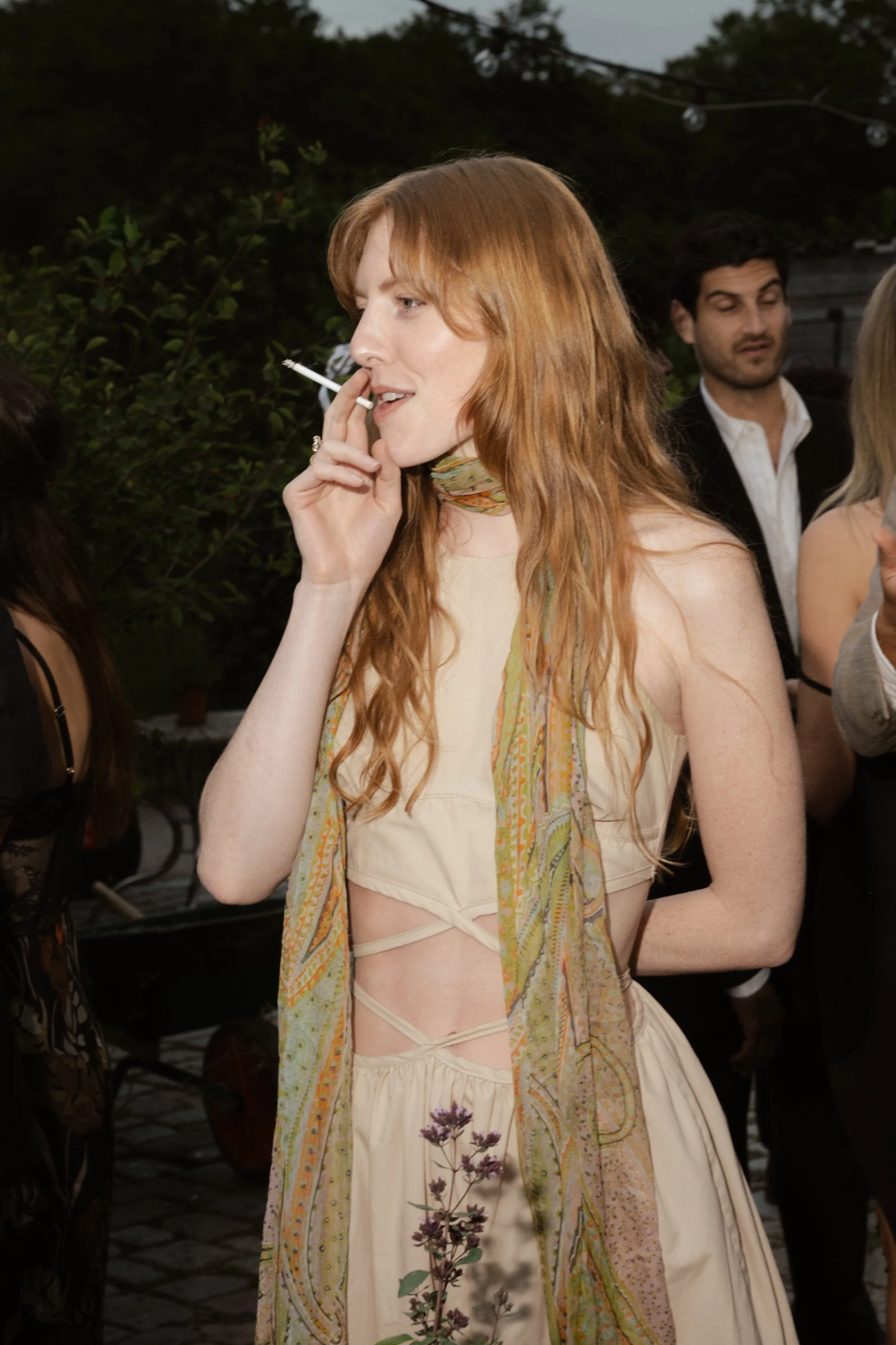 A woman with long red hair wearing a beige dress with cutouts, holding a cigarette, at an outdoor gathering.