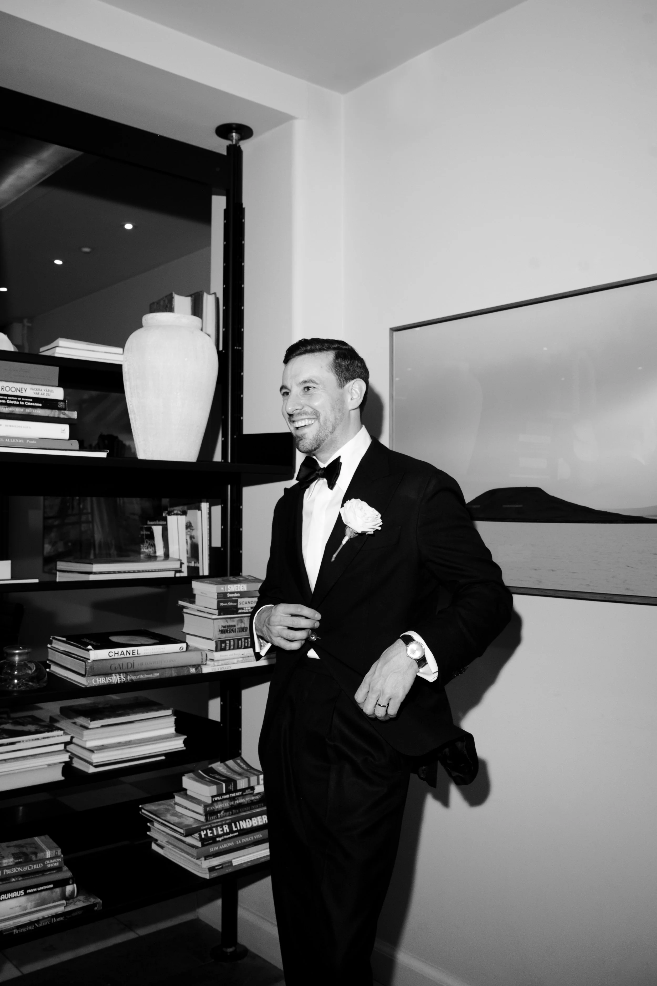 A man in a tuxedo with a boutonnière, standing indoors next to a bookshelf and a framed landscape picture, smiling.