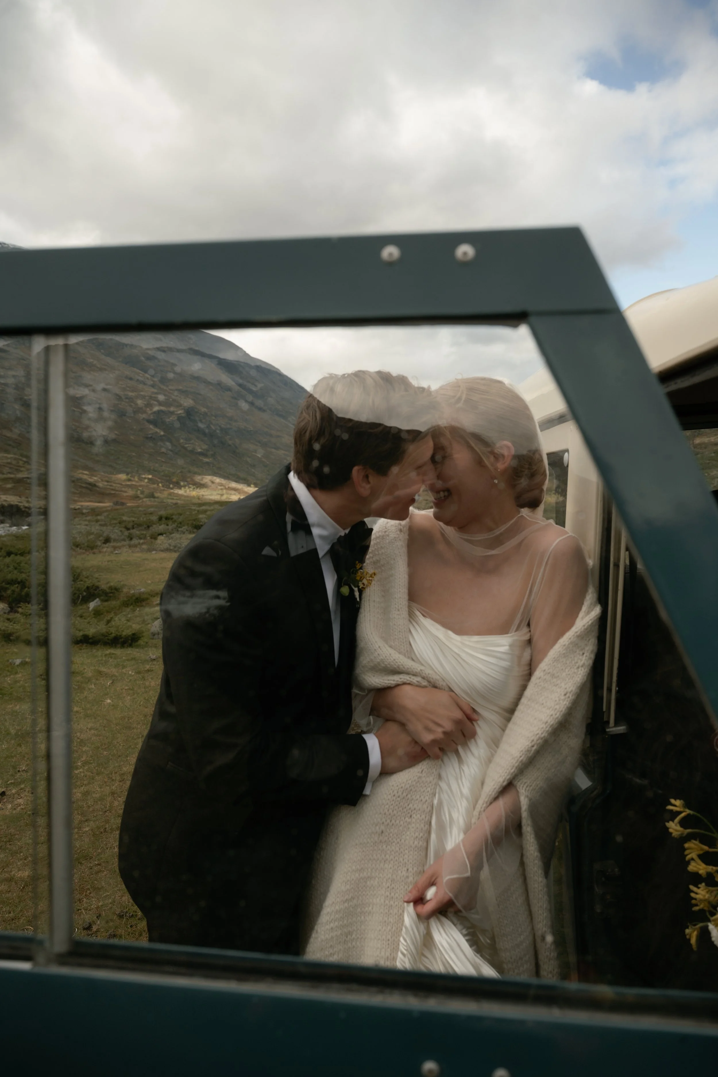 A couple dressed in wedding attire sharing an intimate moment, seen through a window, with a mountain landscape in the background.