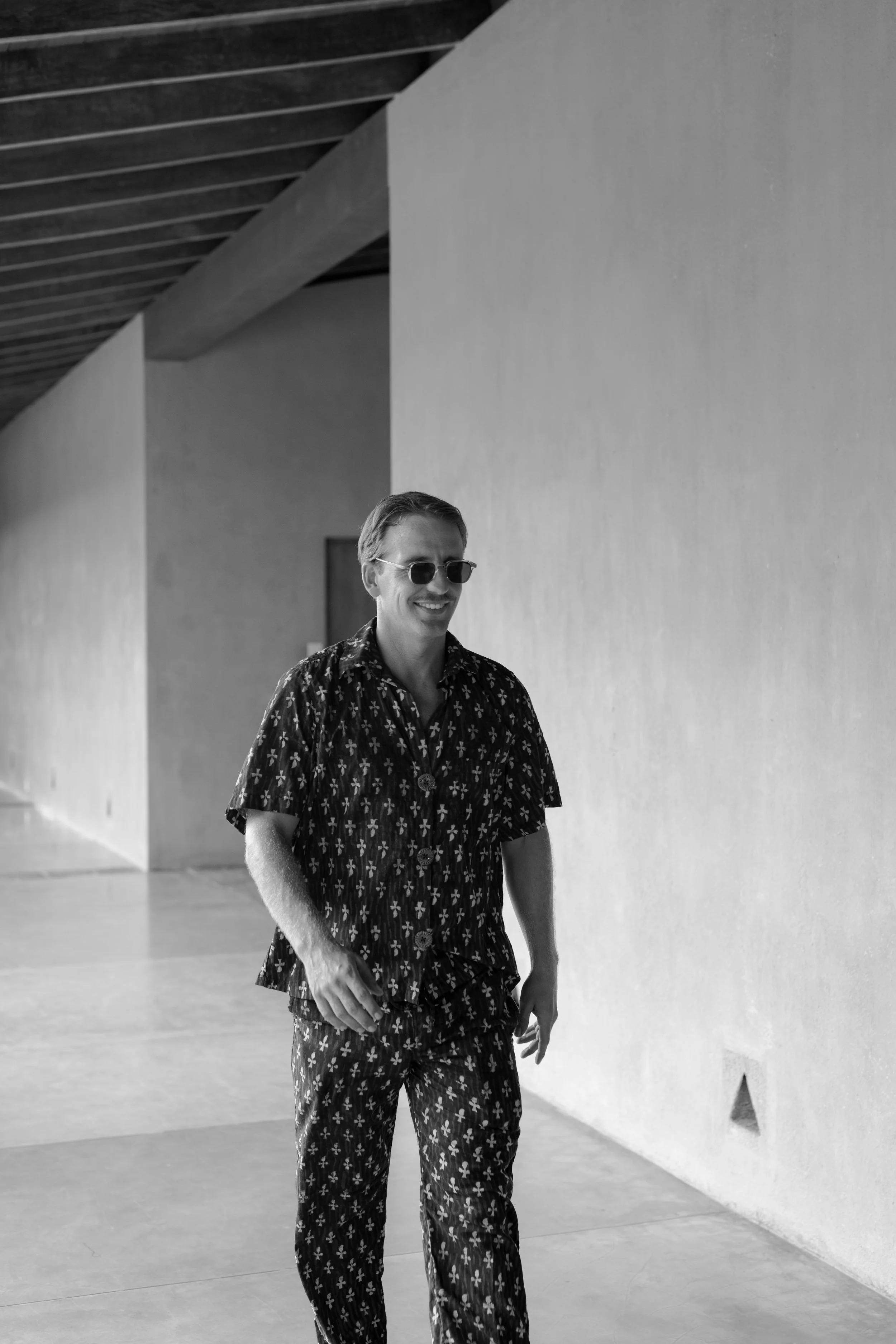 A man walking indoors, wearing sunglasses and a floral print shirt and pants in black and white.