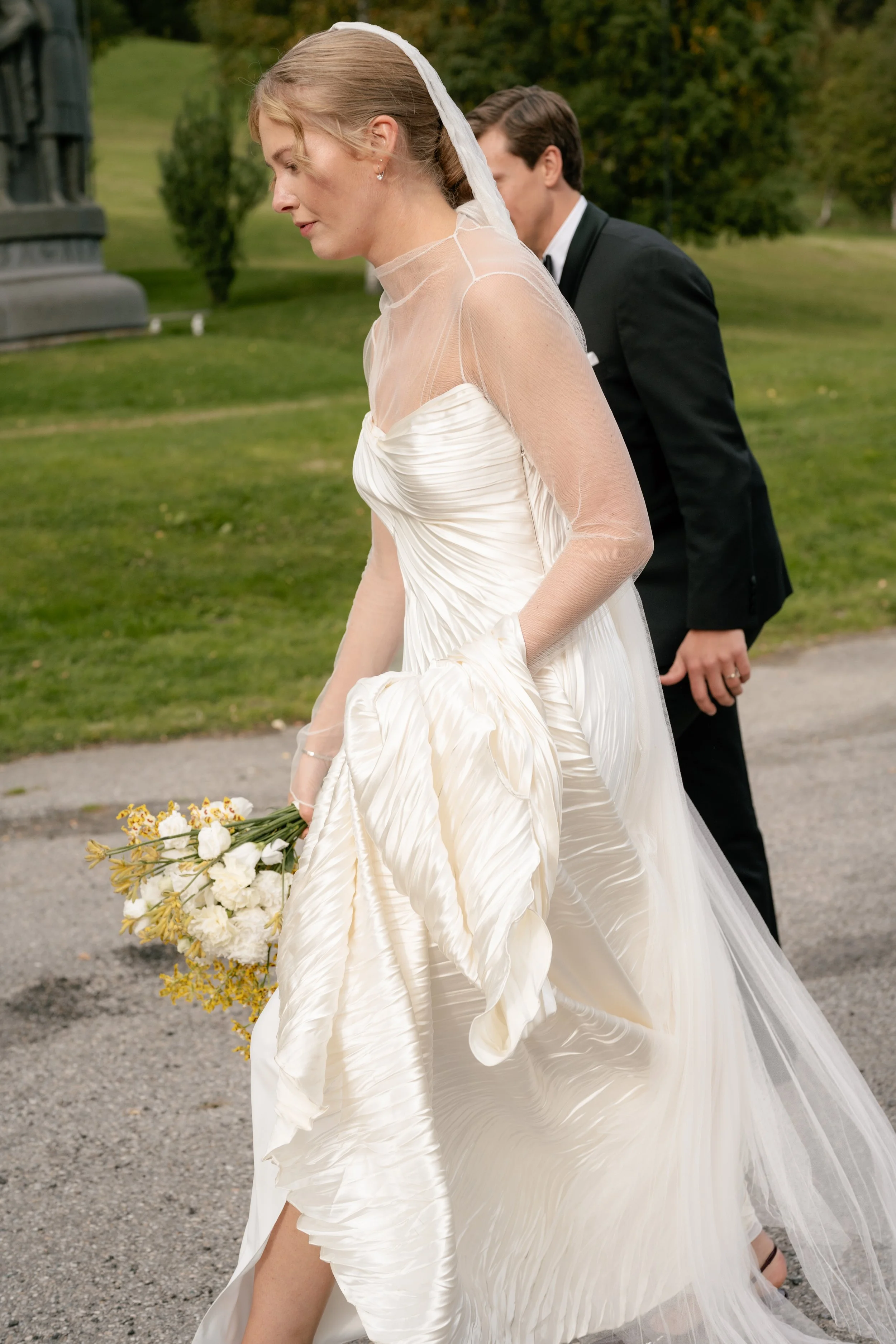 A bride in a white wedding gown holding a bouquet of white flowers and a groom in a black tuxedo standing outside on a pathway in a park-like setting with green grass and trees.