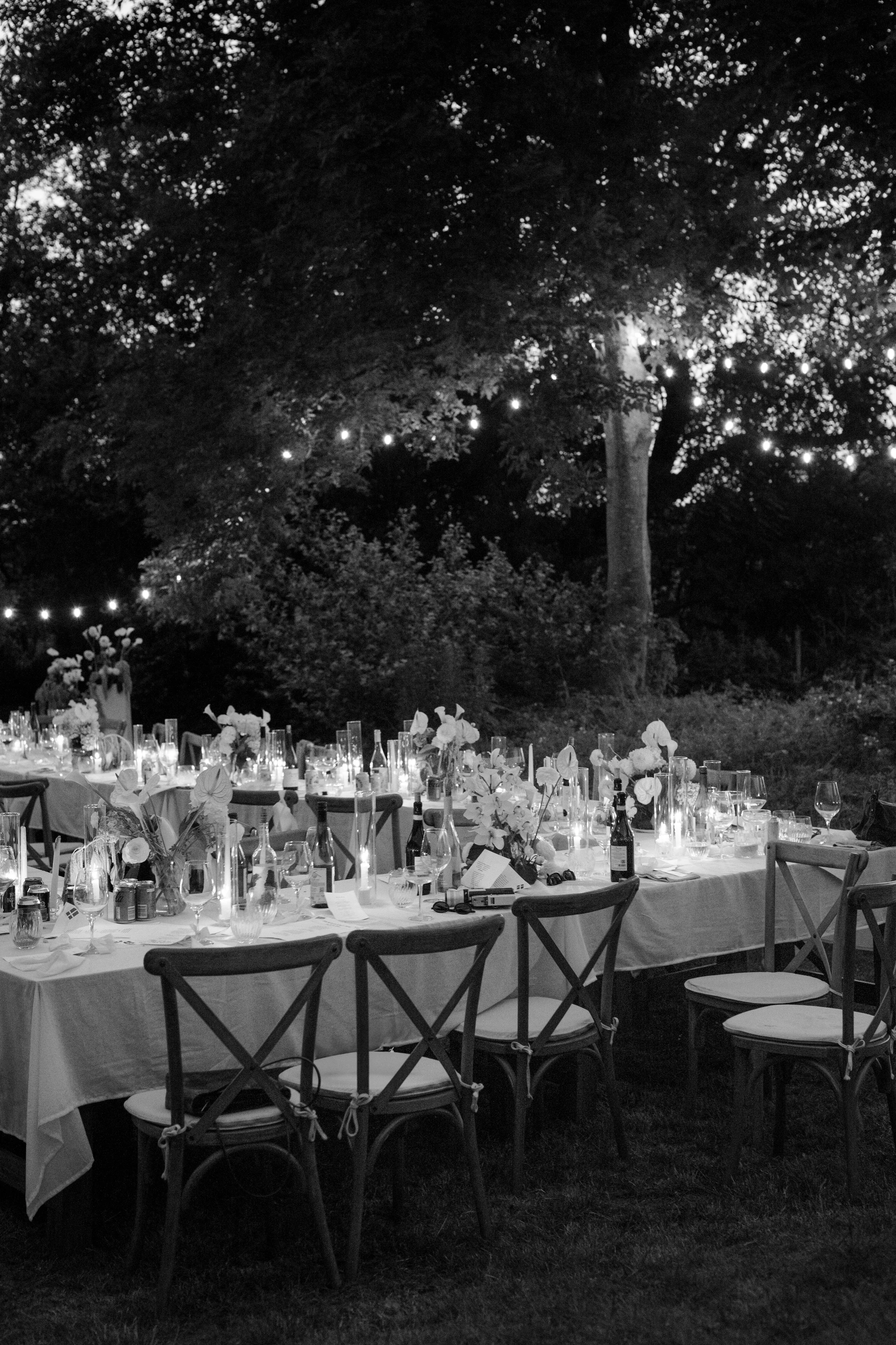 A nighttime outdoor event with decorated tables, chairs, candles, and floral centerpieces set beneath string lights in a wooded area.