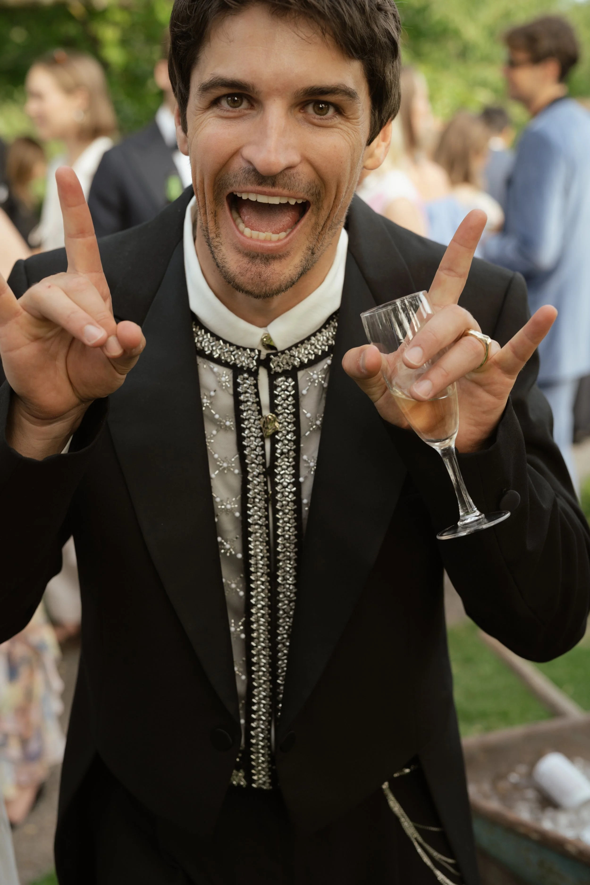 Man smiling and making a 'rock on' gesture at an outdoor celebration, holding a glass of champagne, wearing a black suit with a sequined shirt.
