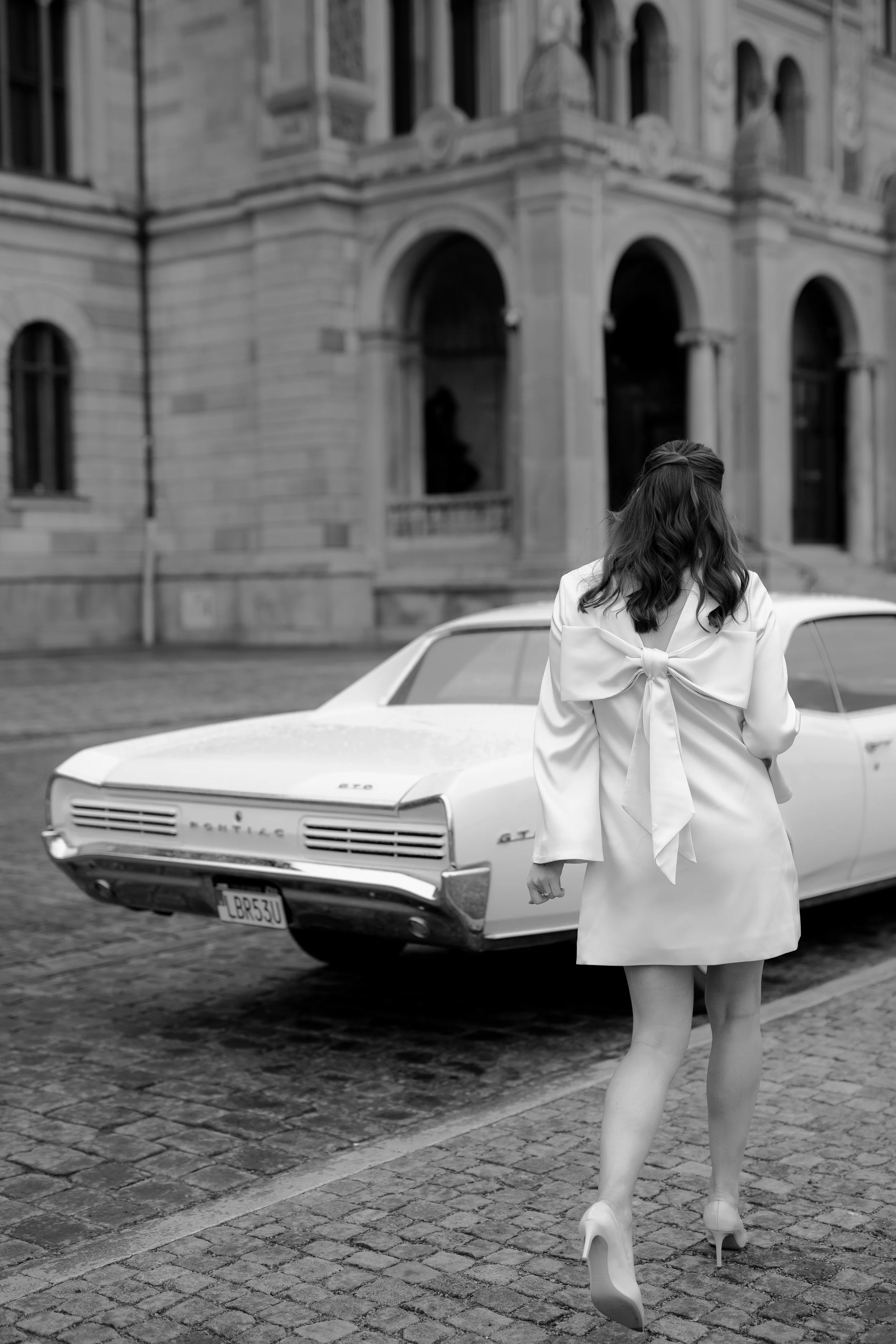 A woman in a white coat with a large bow on the back, wearing high heels, walks away from a vintage car on a cobblestone street in front of a historic building.