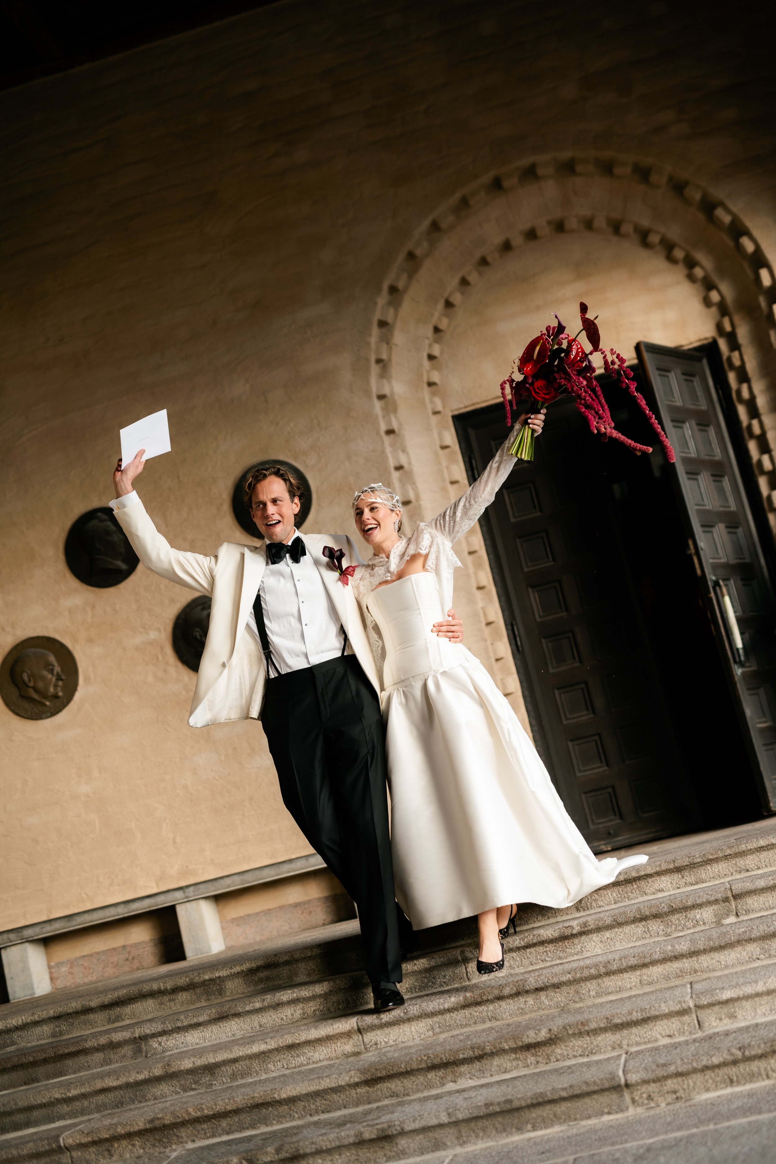 A bride and groom celebrating on the steps of a building, with the bride holding a bouquet and the groom raising a card, both smiling.