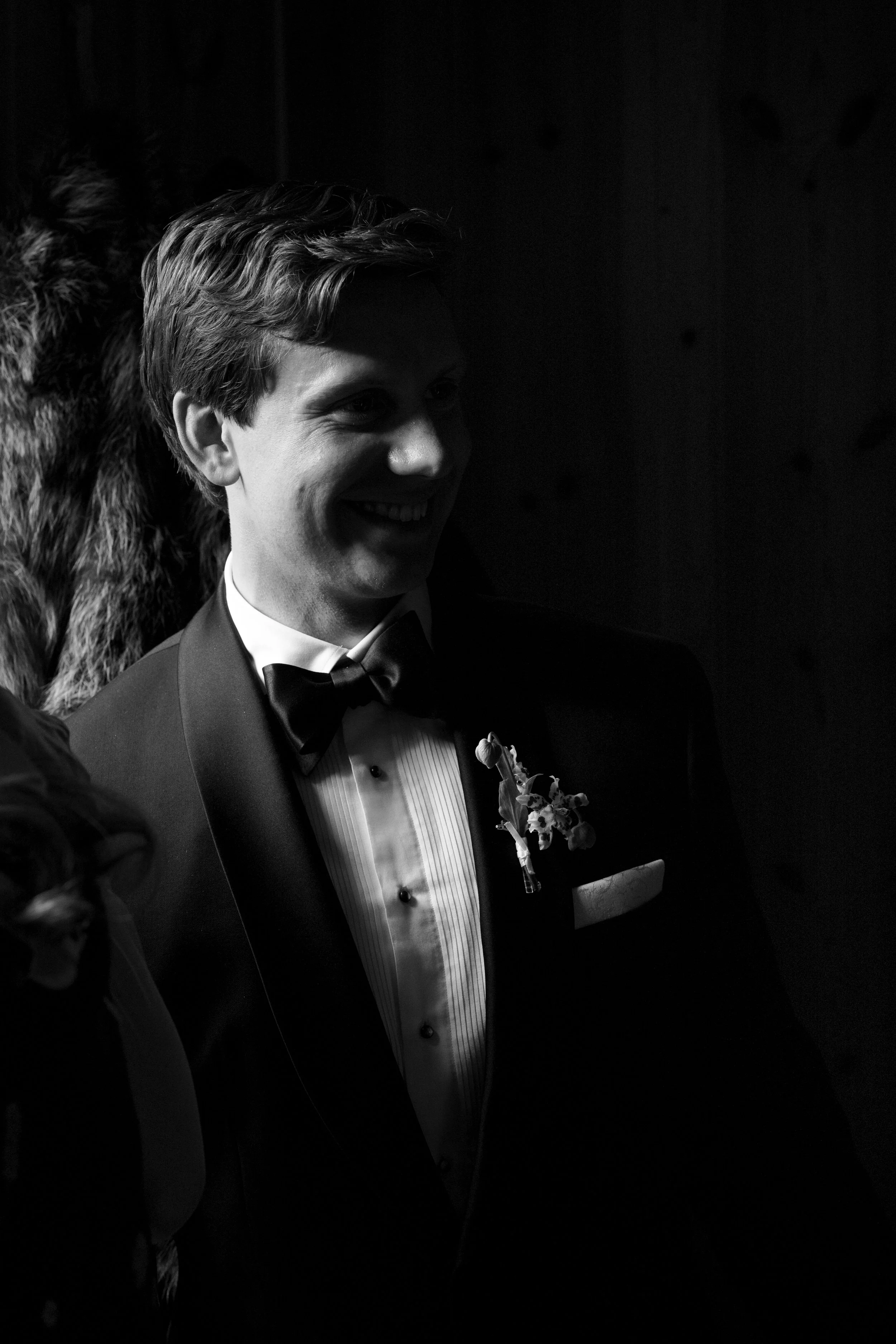 A young man in formal tuxedo with a bow tie, smiling, standing against a dark background with a fur coat draped behind him.